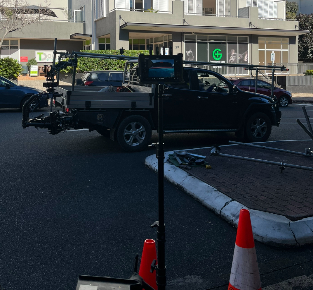 A black utility truck fitted with camera and drone equipment parked on the street in front of a building with a green veterinary clinic sign reading "Greencross Vets". Two orange safety cones are in the foreground, and various equipment is on the ground nearby.