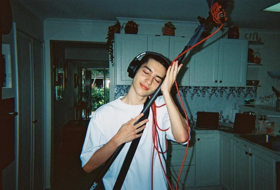 Young man with short dark hair wearing headphones, holding a microphone stand with red cables, smiling with eyes closed in a kitchen.