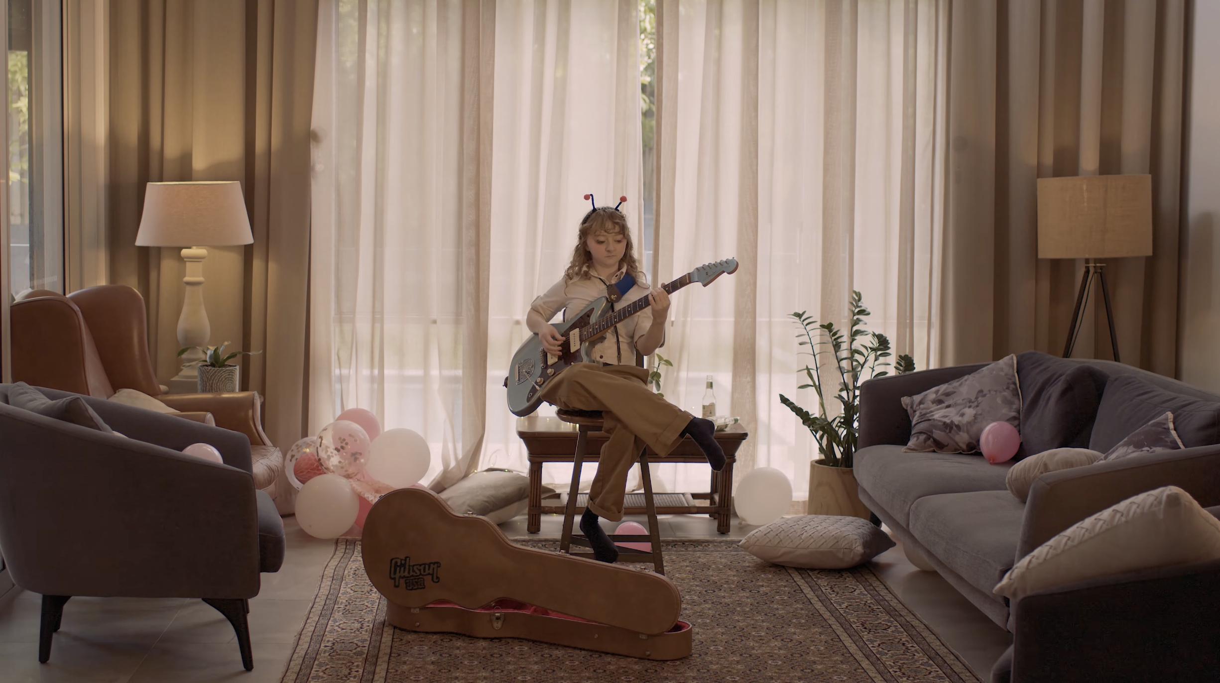 A young girl with curly hair wearing a white shirt and brown pants, sitting on a stool in a living room, playing an electric guitar. There are balloons and decorations on the floor, a couch on the right, armchairs on the left, and a large window with curtains behind her, with sunlight filtering through.