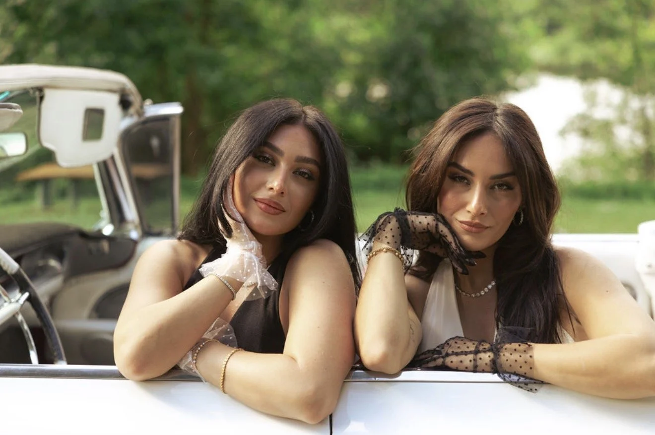 Two women with dark hair leaning on a white vintage car, smiling at the camera outdoors with green trees in the background.