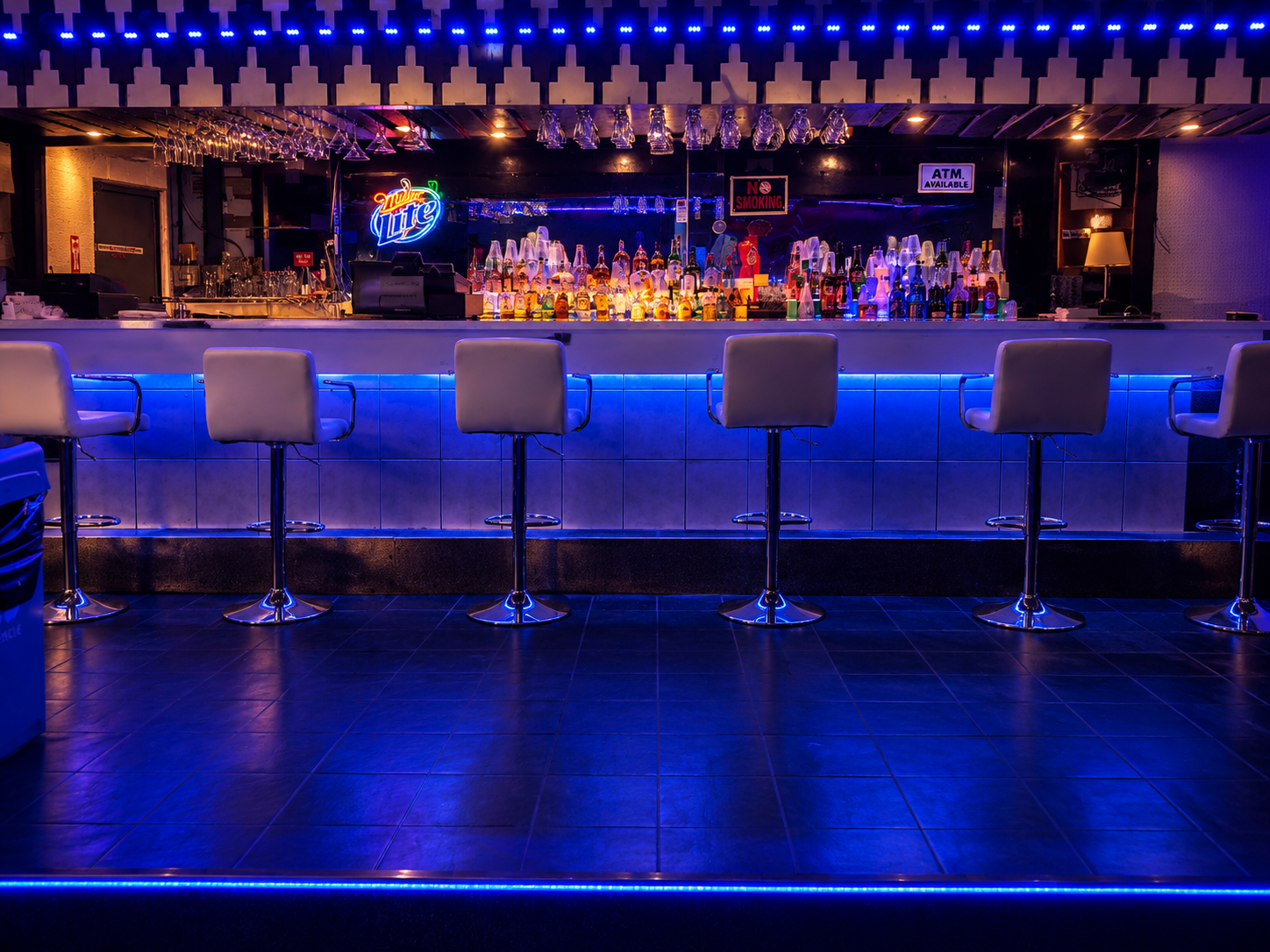 Empty bar with white bar stools, illuminated with blue neon lights, bottles of alcohol on the shelves, neon signs, glasses hanging upside down, and signs displaying 'No Smoking' and 'ATM Available'.