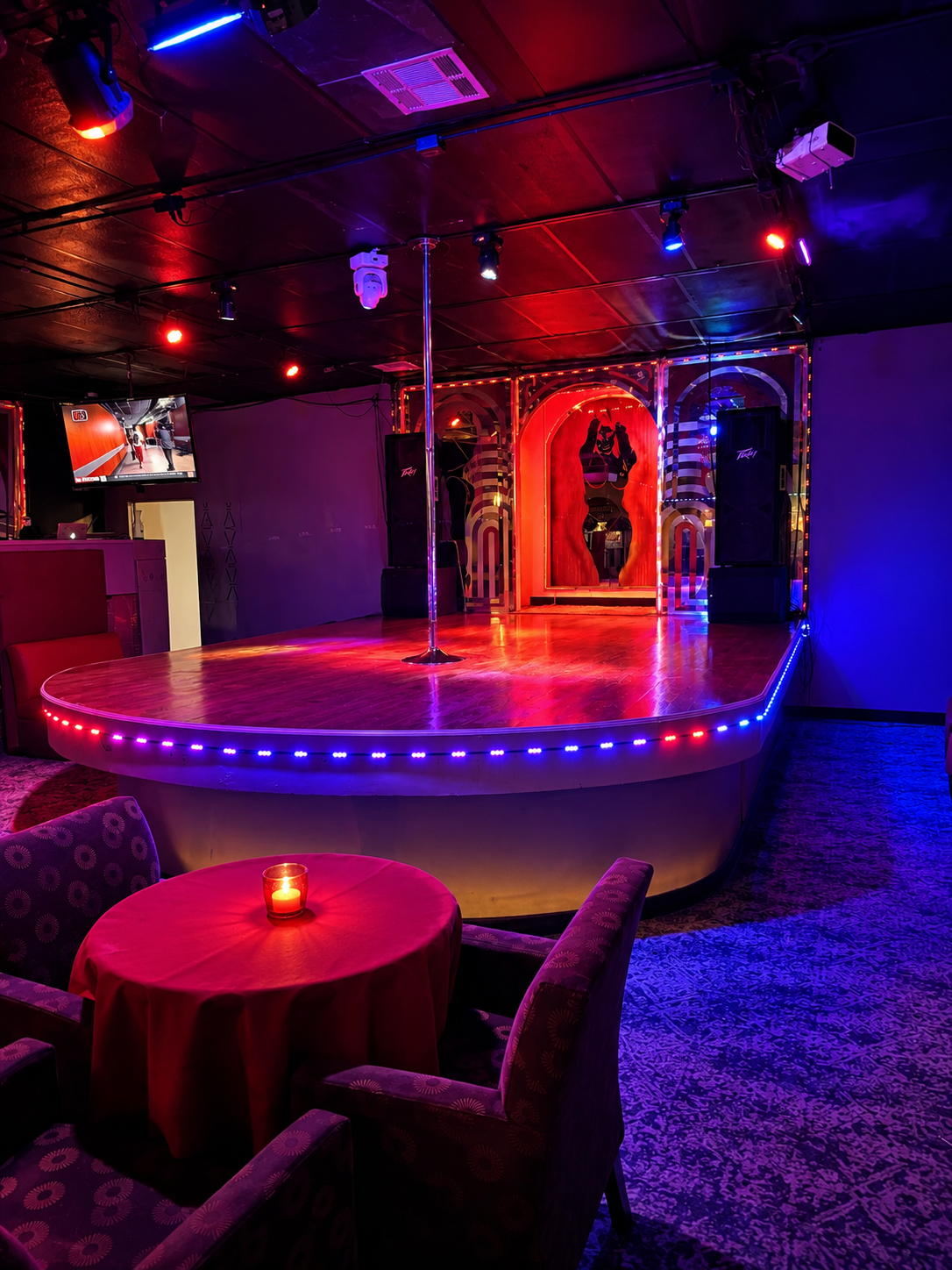A small stage with a wooden floor and a lone pole in the center, illuminated by colorful red and blue LED lights. Behind the stage is a decorative mirror with neon lights, a silhouette of a woman, and striped arches. To the left, a TV screen displays a corridor scene, with a small table and chairs nearby, lit by a candle.