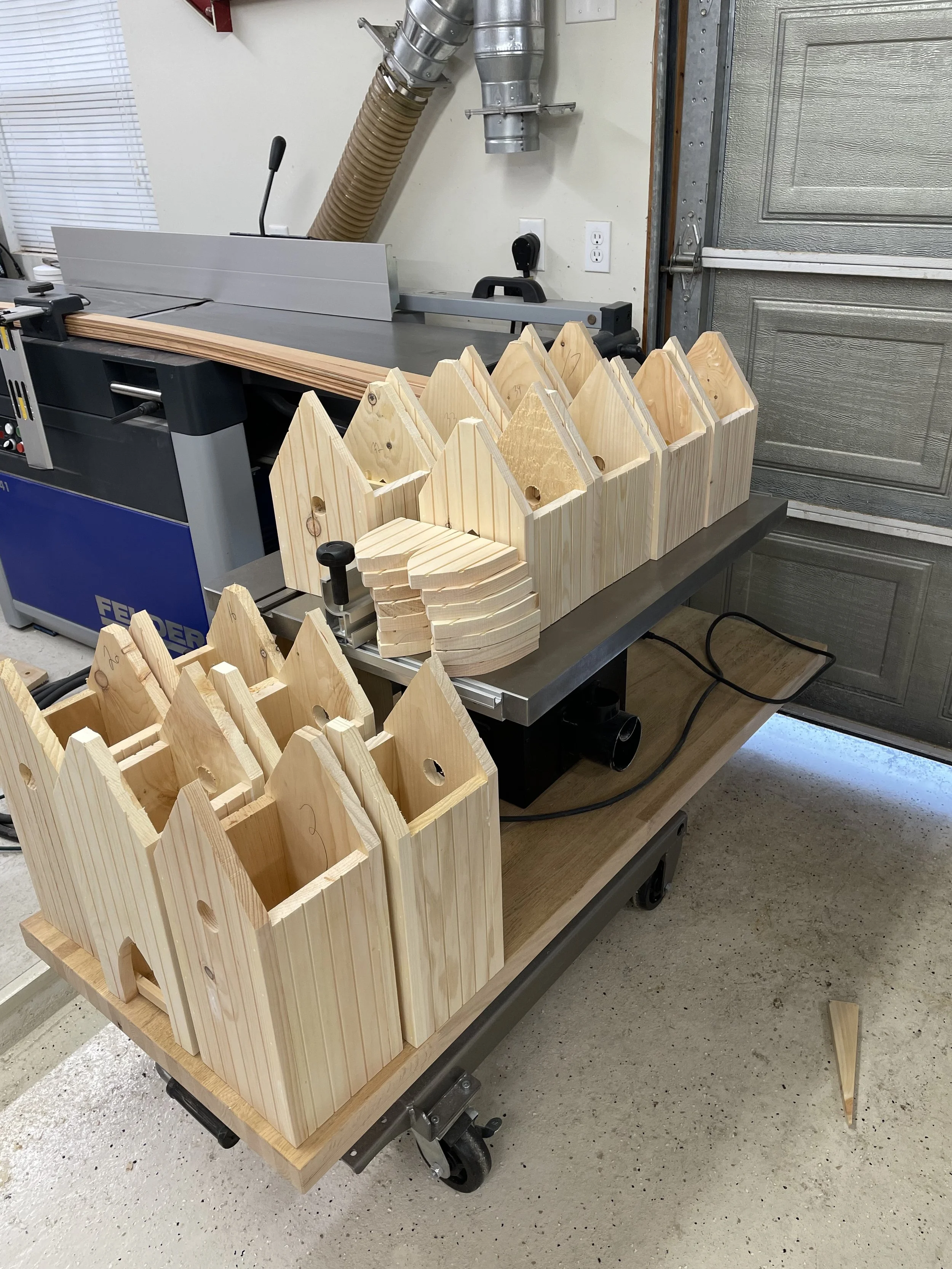 Wooden birdhouses in various stages of construction on a workbench with a table in a garage or workshop area.