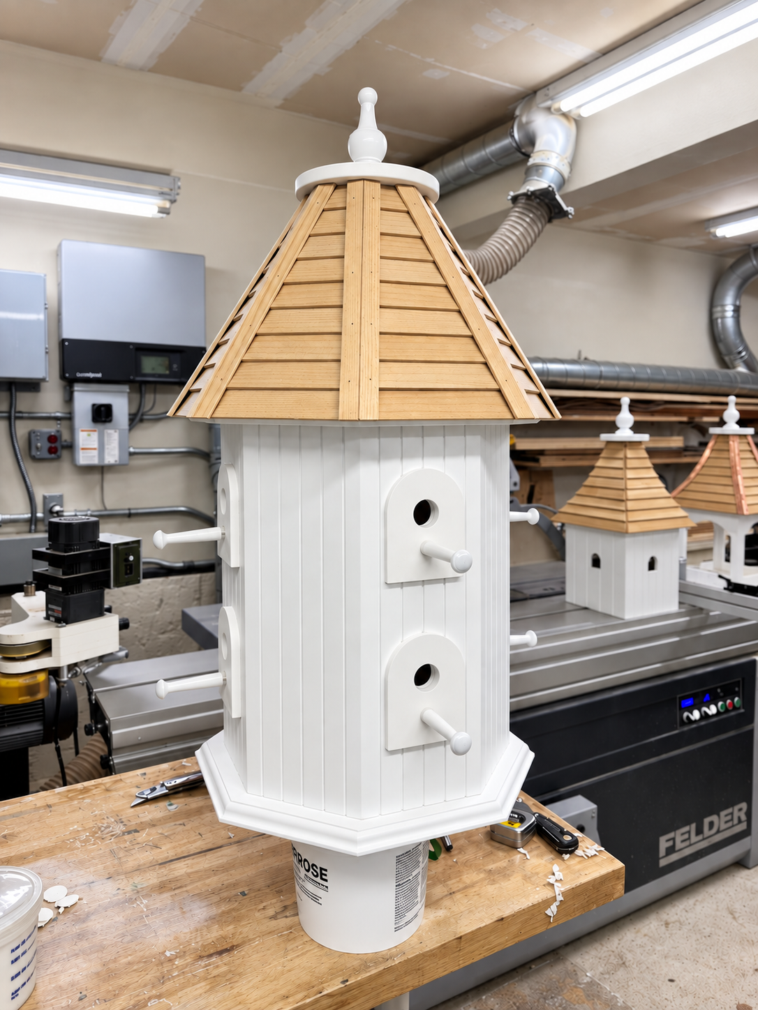 A woodworking workshop with a completed birdhouse featuring a white base, vertical siding, and a brown shingled roof, placed on a workbench.