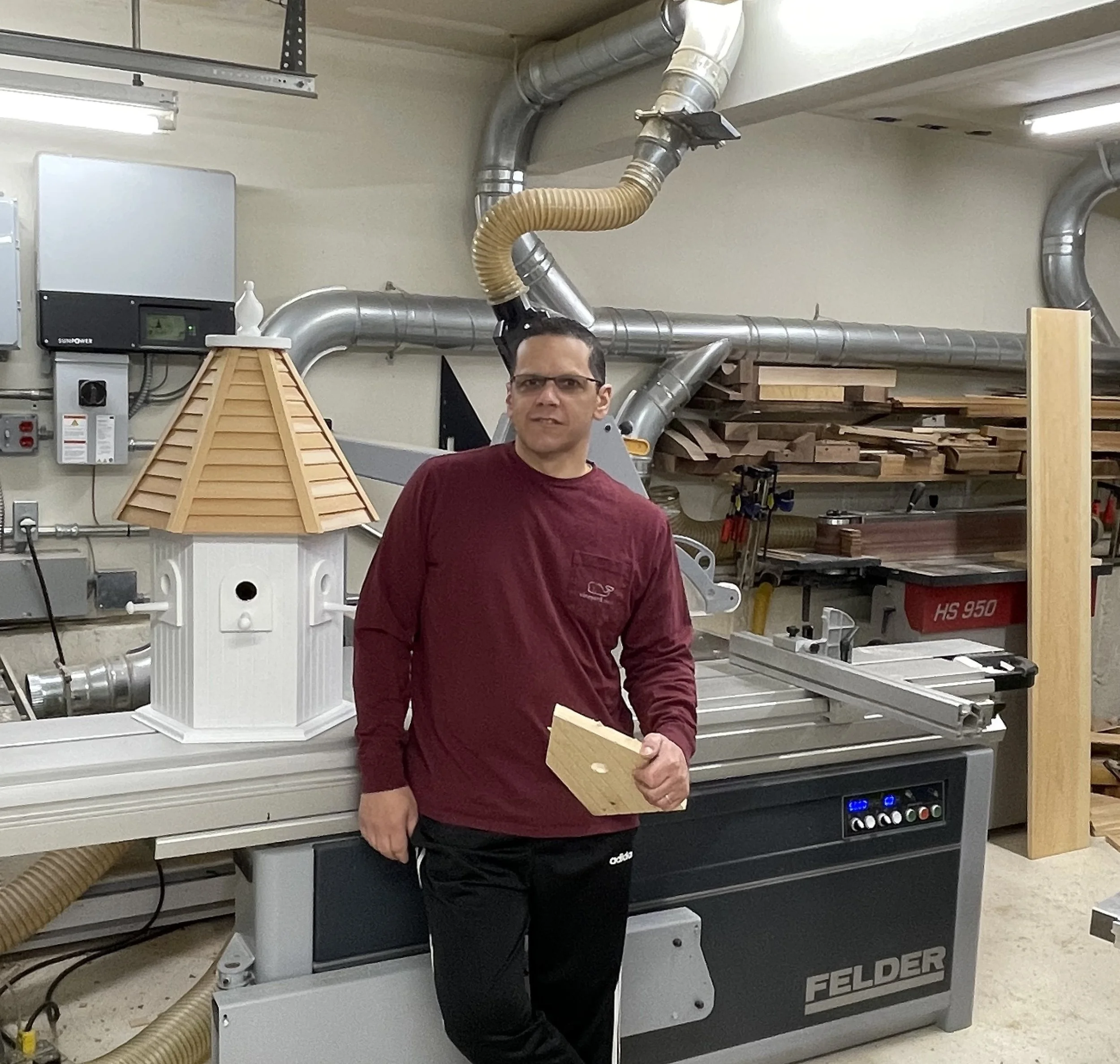A man in a maroon shirt and black pants standing in a woodworking workshop, holding a piece of wood, with wood and machinery around him.