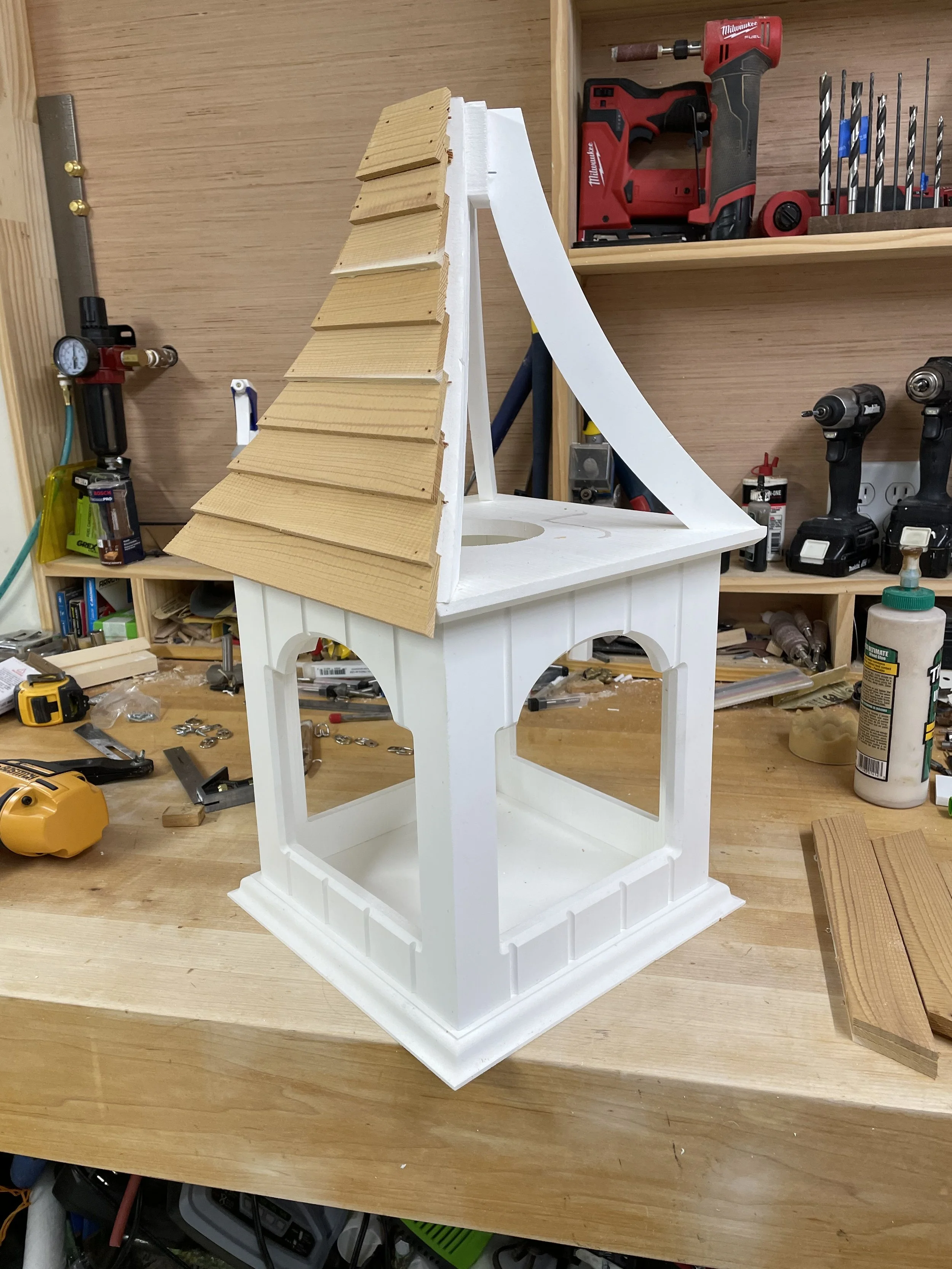 A partially assembled white bird feeder on a workbench, with tools, drill bits, and woodworking supplies in the background.
