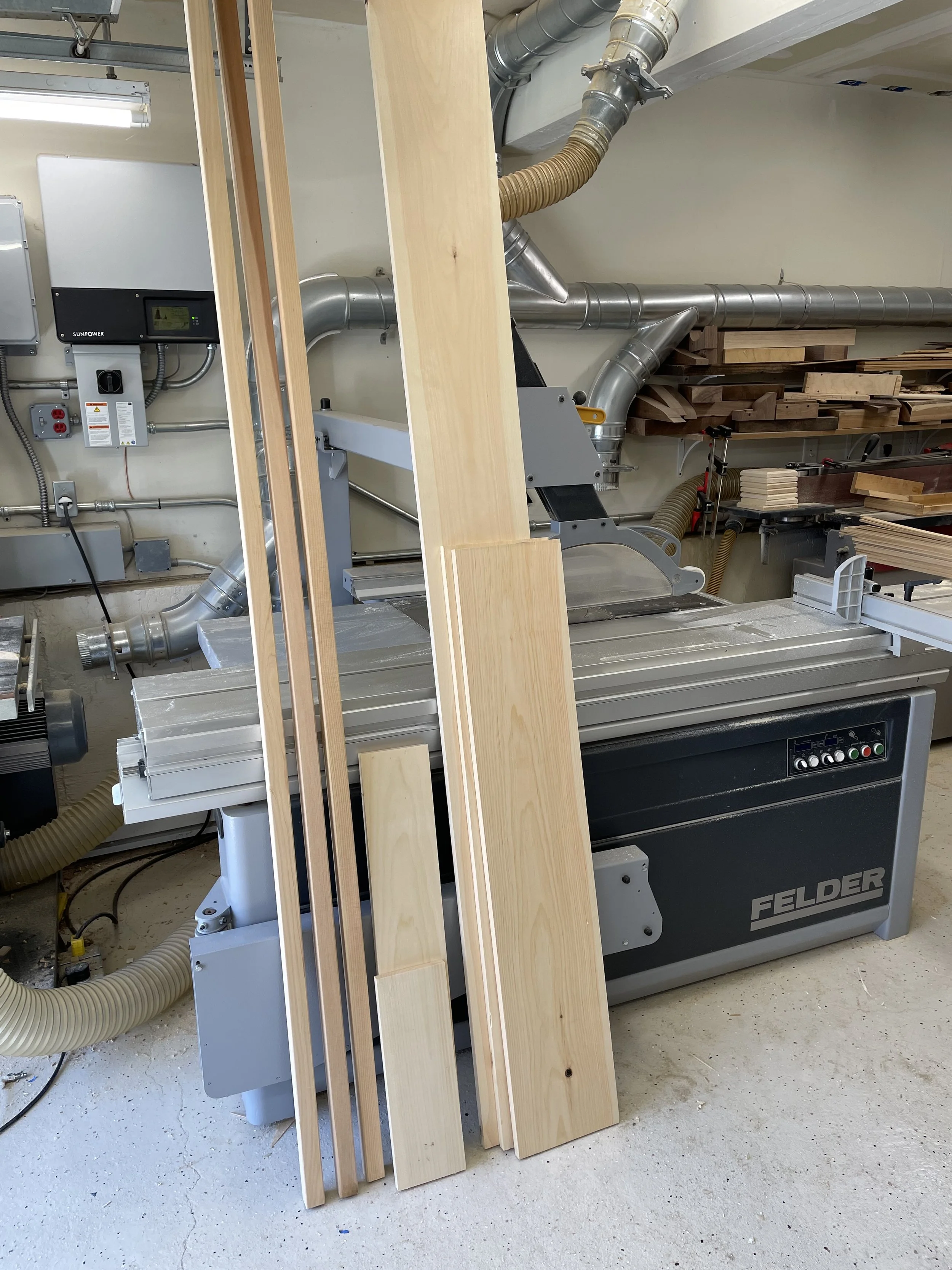A woodworking shop with a Felder machine, several unfinished wooden panels, and sawdust on the floor.