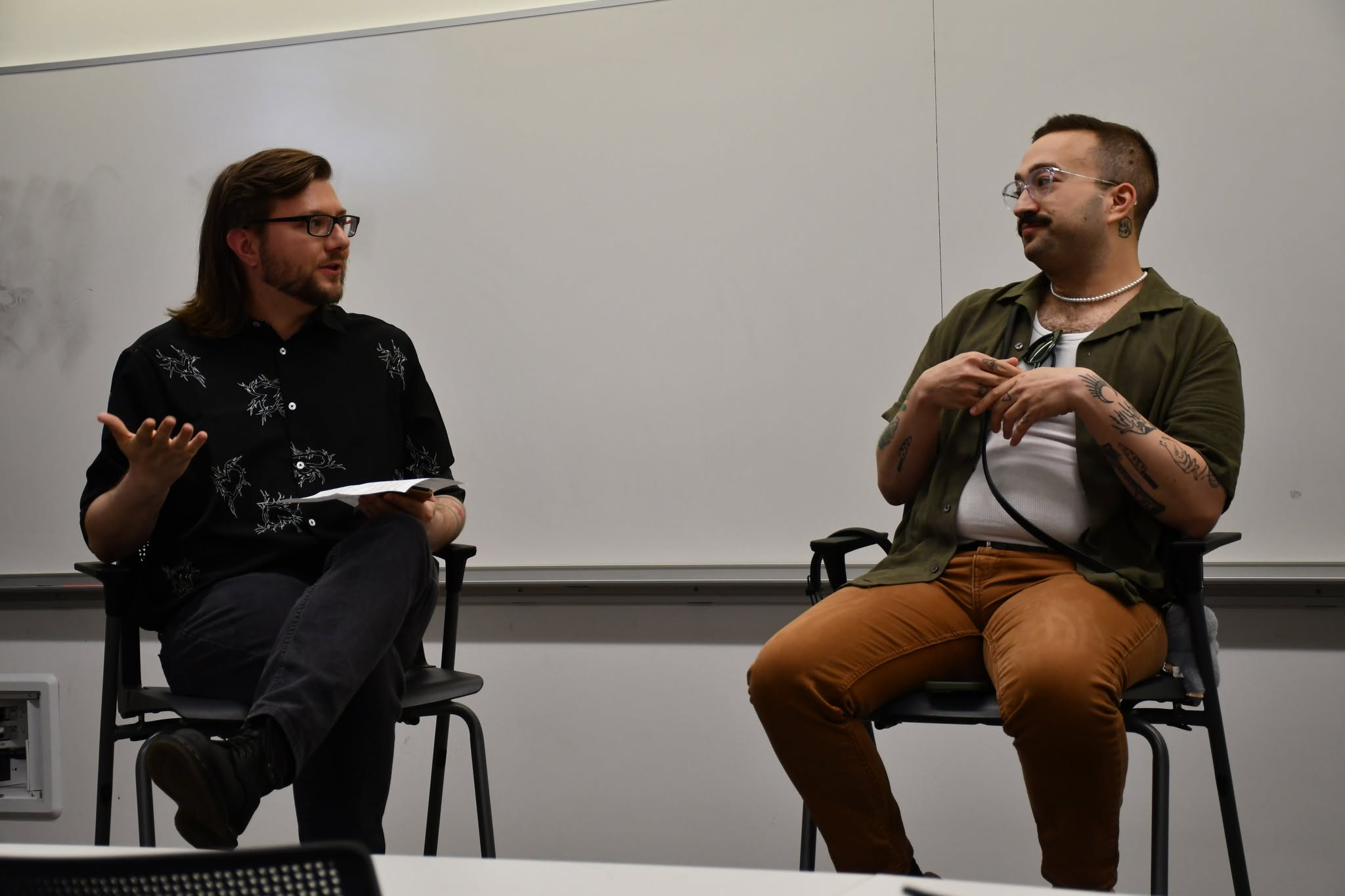 Jake Wittich and Adam Rhodes sitting and talking in a classroom or lecture hall, Jake is gesturing with a hand while holding papers, Adam with tattoos and glasses listening.
