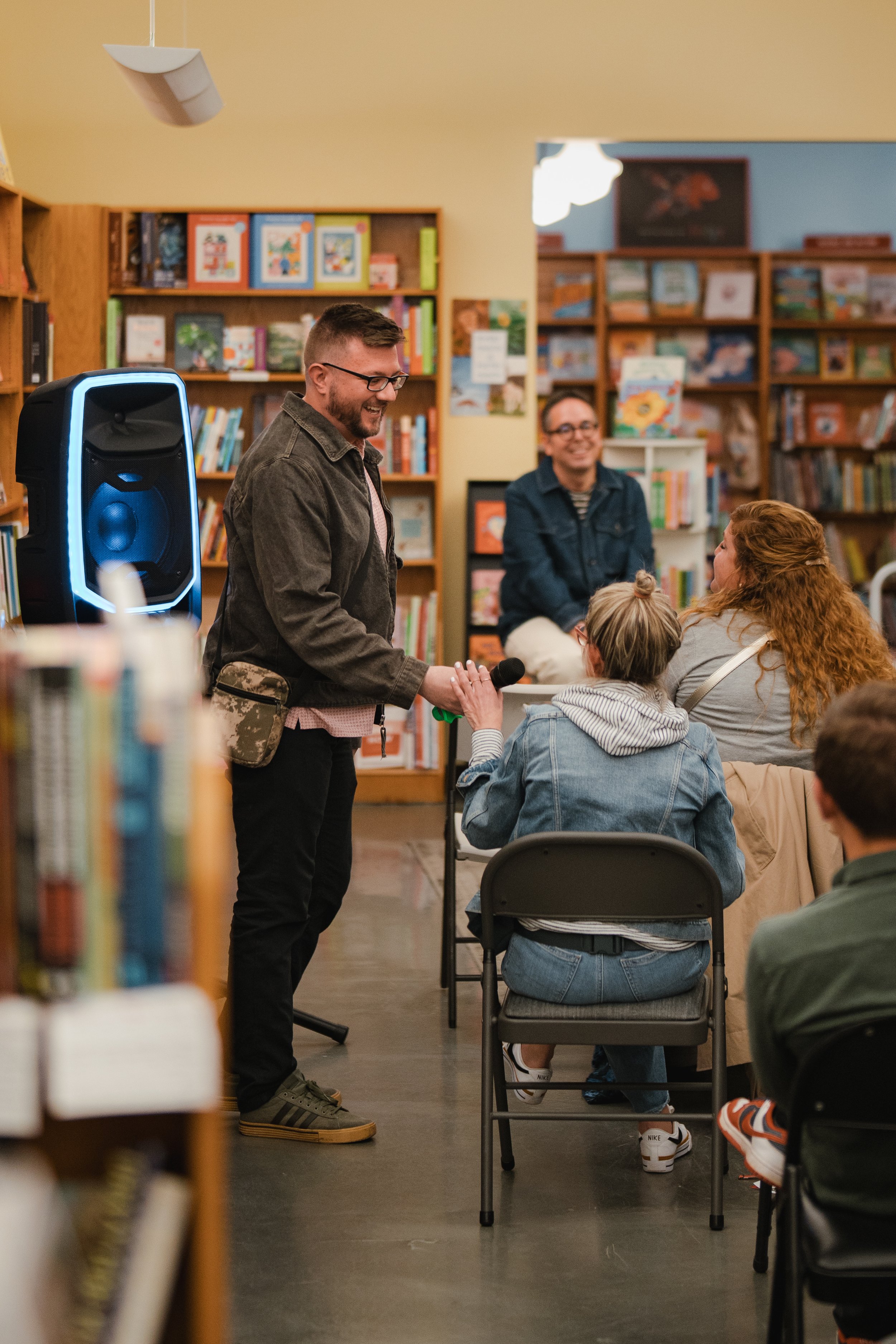 Jake Wittich smiling and holding a microphone, engaging with a woman seated at a table in a bookstore or library. Behind him, two other people are seated, and there are bookshelves filled with colorful books in the background.