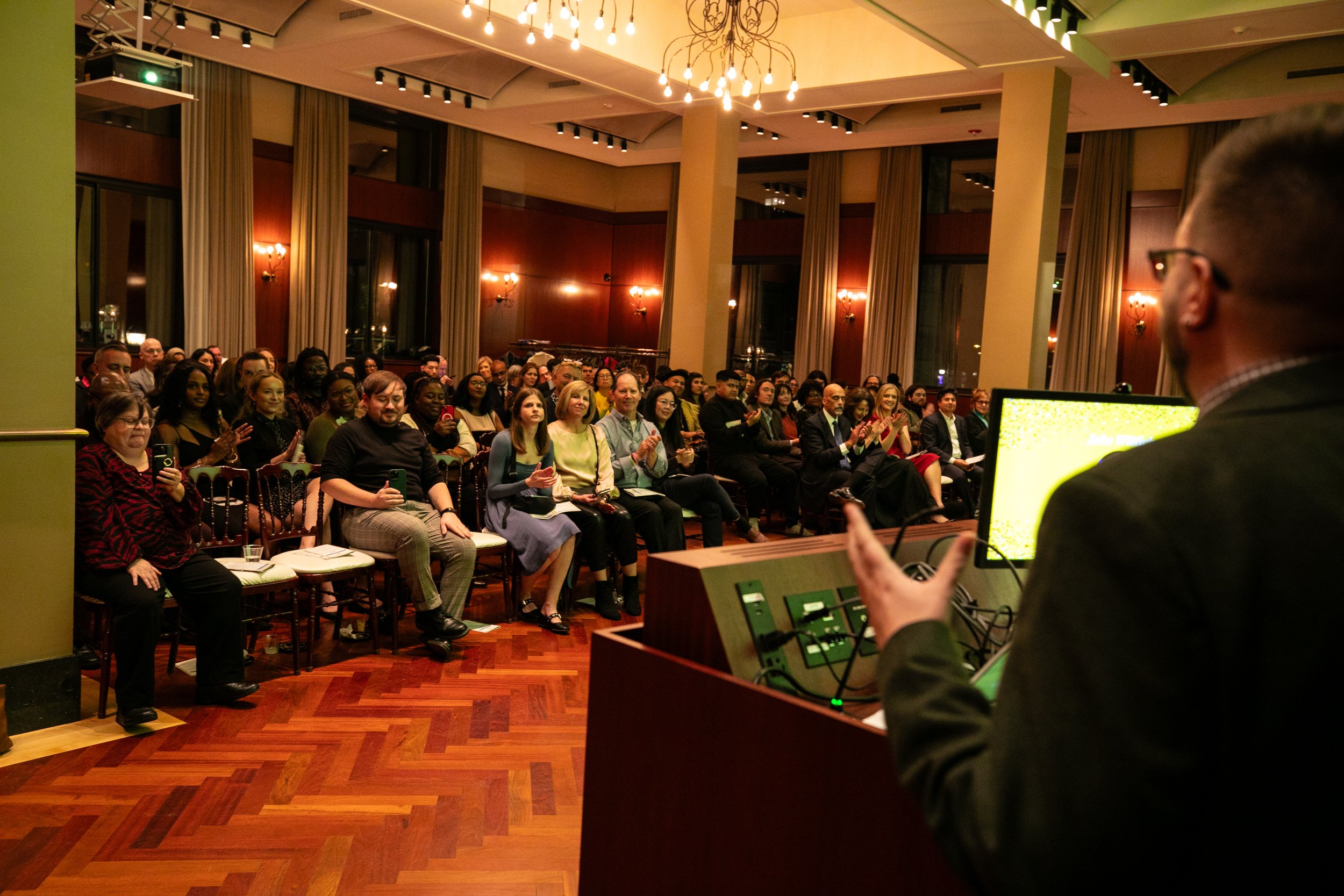 Jake Wittich speaks to A large group of diverse people seated in a conference room.