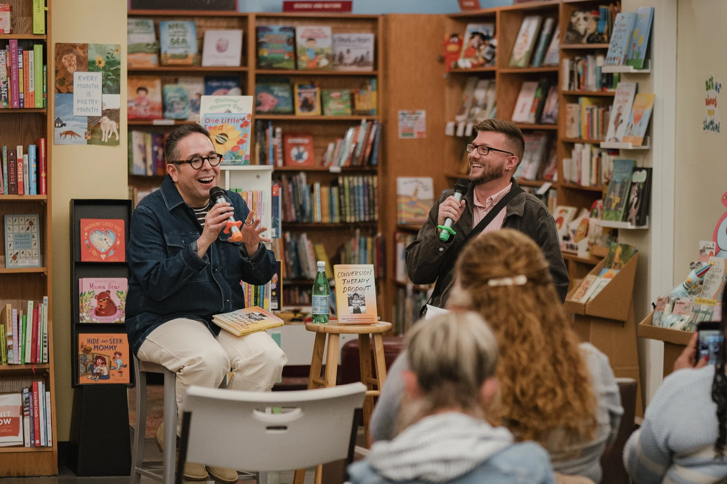 Timothy Schraeder Rodriguez and Jake Wittich sitting and speaking with microphones in a bookstore event surrounded by shelves filled with children's books and posters, engaging with an audience.