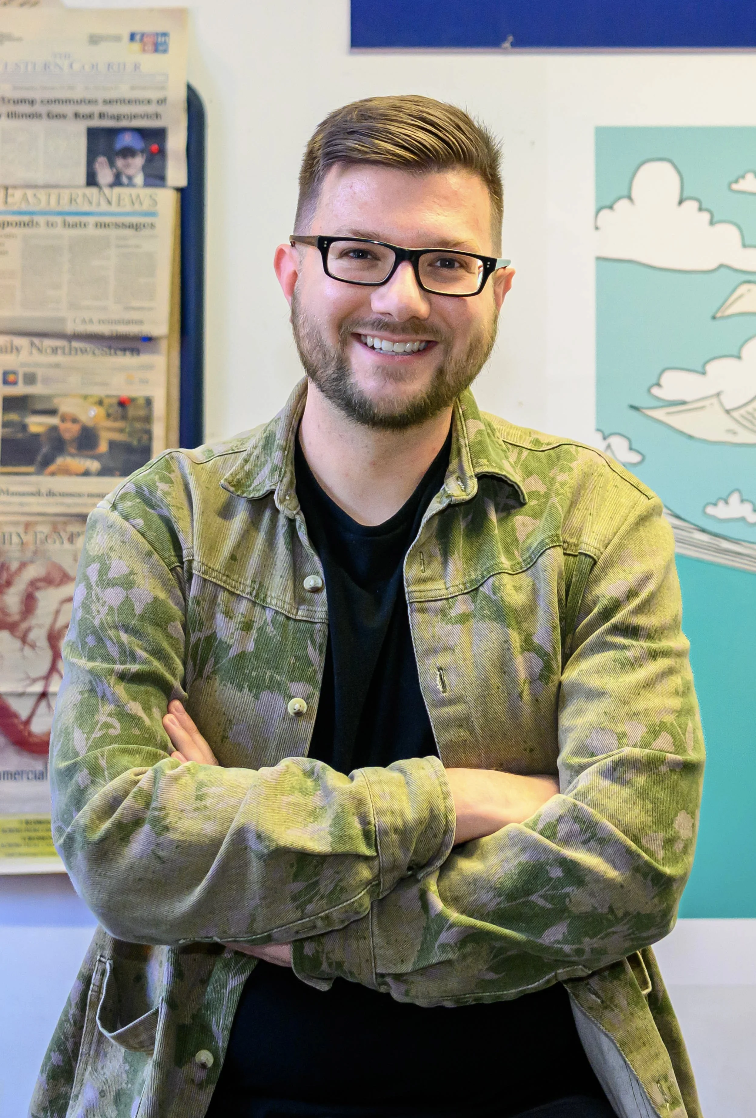 Jake Wittich smiling, wearing a green camouflage-patterned jacket with a black shirt underneath, standing with arms crossed in front of a blue and white wall with posters and a cartoonish depiction of clouds and paper airplanes.