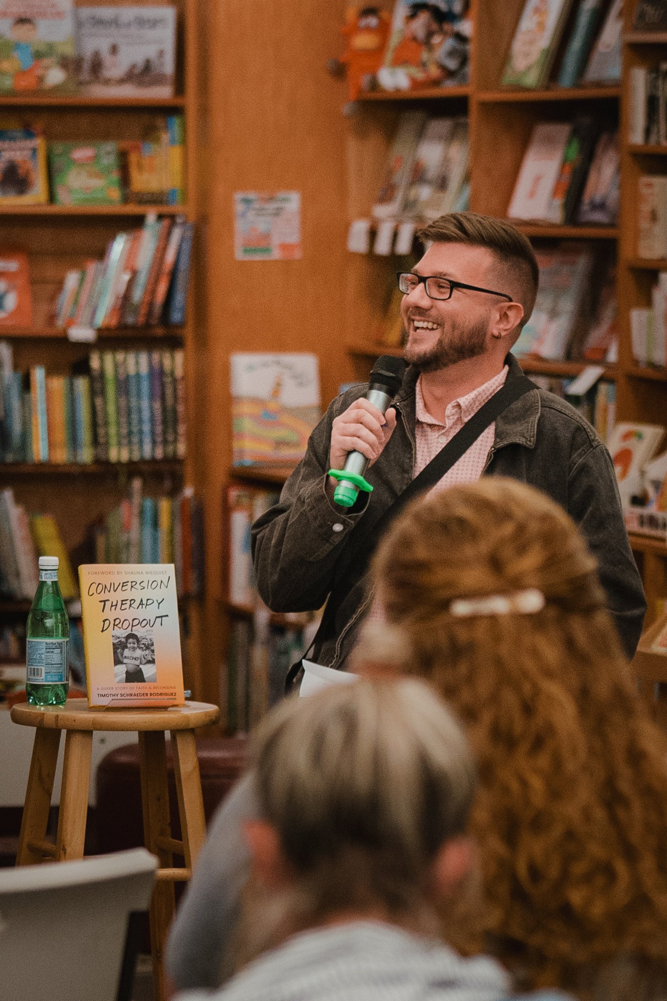 Jake Wittich holding a microphone, smiling, in a bookstore or library setting with shelves of books in the background.
