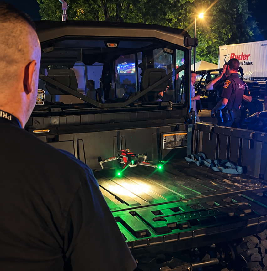 A police officer is standing near a black utility vehicle with a drone on its cargo area, illuminated by green drone lights, during nighttime with emergency personnel in the background.