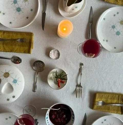 A top-down view of a holiday dinner table with plates, silverware, glasses of red wine, candles, and a small bowl with a sprig of holly. There is a white tablecloth and a dish with cranberry sauce.