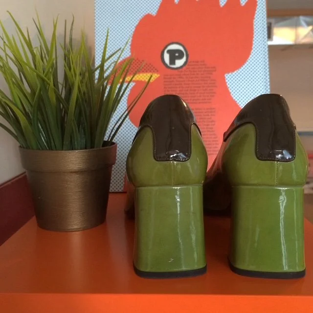 Back view of a pair of green and black platform shoes on an orange table, with a potted plant and a colorful poster in the background.