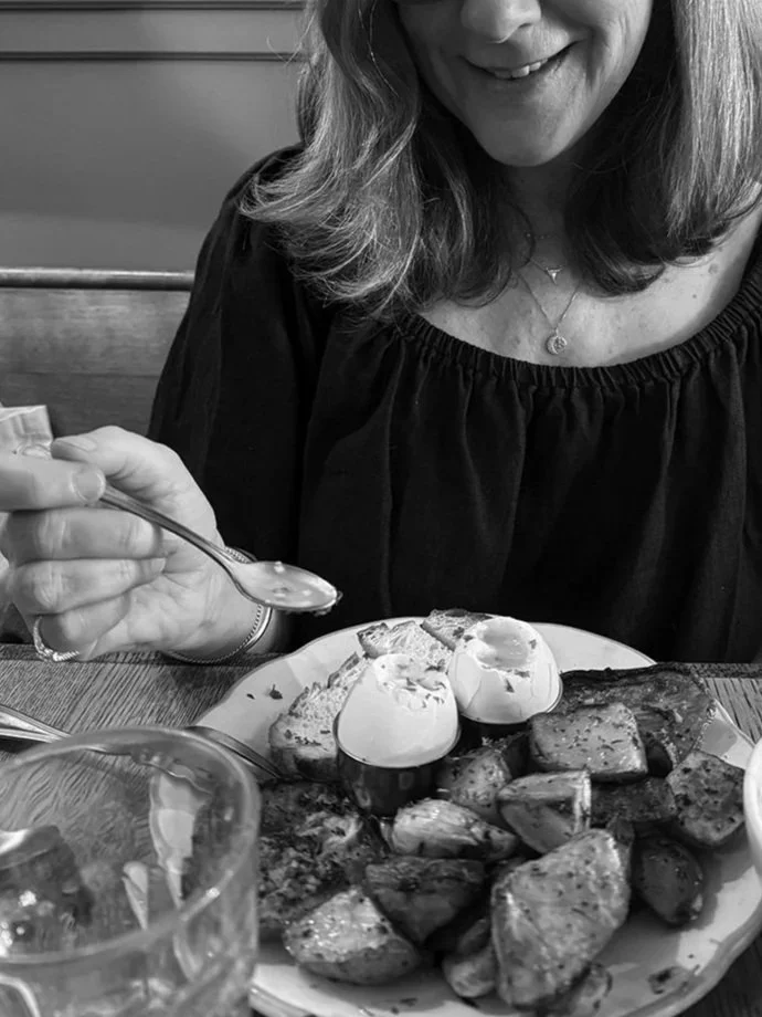 A woman with shoulder-length hair, wearing a black top and a necklace, is holding a spoon above a plate of baked potatoes and two soft-boiled eggs. The image is in black and white.