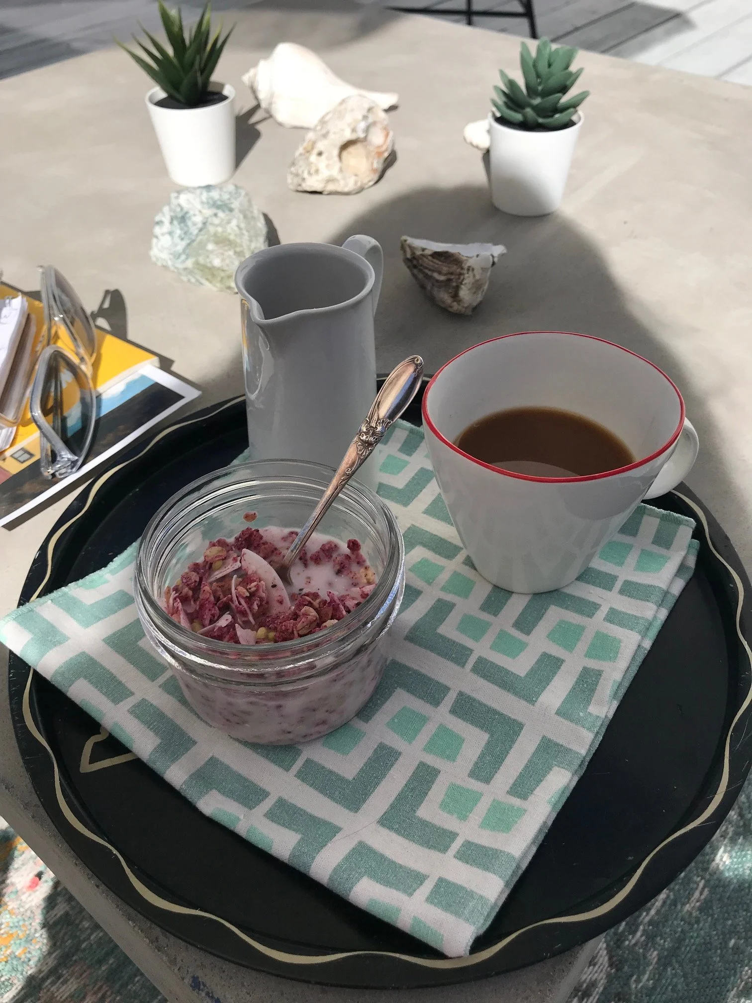 A tray with a glass jar of pink yogurt with granola and berries, a white pitcher, a coffee cup filled with coffee, a pair of sunglasses, and some scattered papers on a table with a green and white patterned cloth. In the background, there are two potted succulents and some seashells.