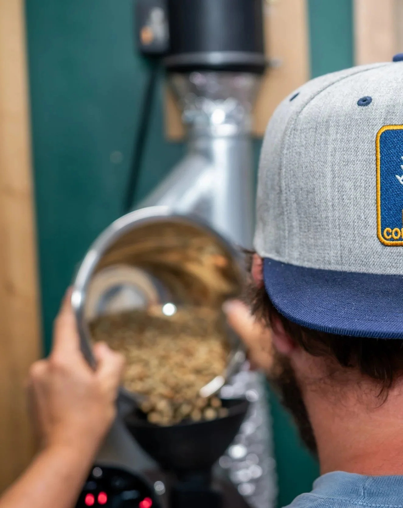 Child wearing a gray and blue cap with an embroidered logo, holding a metal cup, with a coffee maker in the background