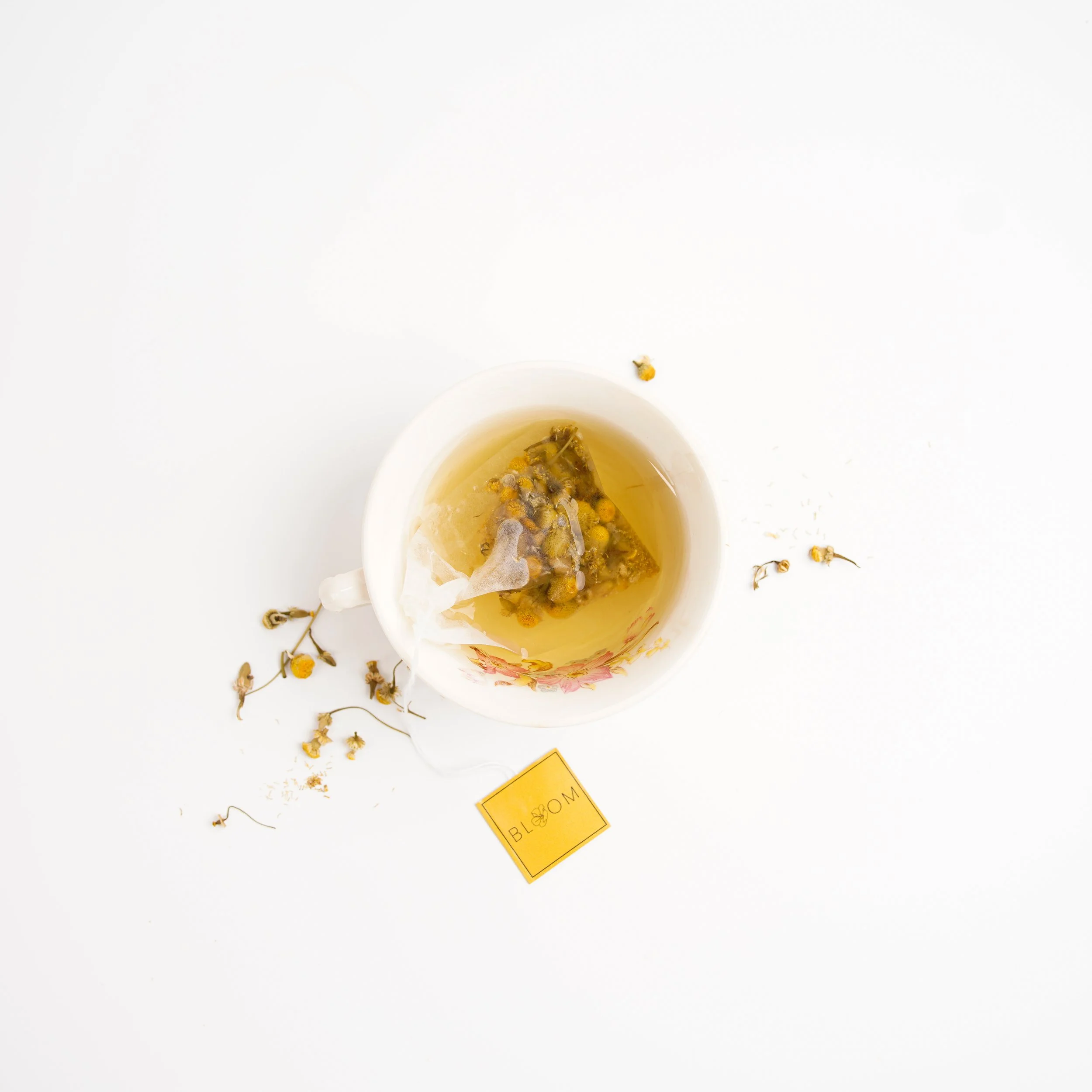 White cup of chamomile tea with dried chamomile flowers inside, placed on a white surface with loose dried chamomile flowers scattered around and a small yellow tag with the word 'Blog' written on it.