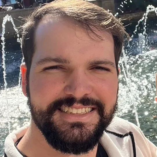 A man with a beard and short hair smiling outdoors near a water fountain.