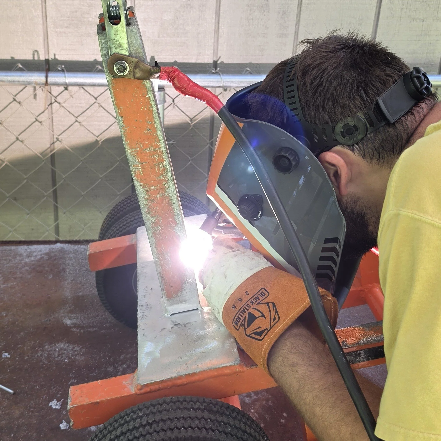 A person wearing safety goggles welding a metal pipe on a construction cart in an outdoor setting with a chain-link fence in the background.