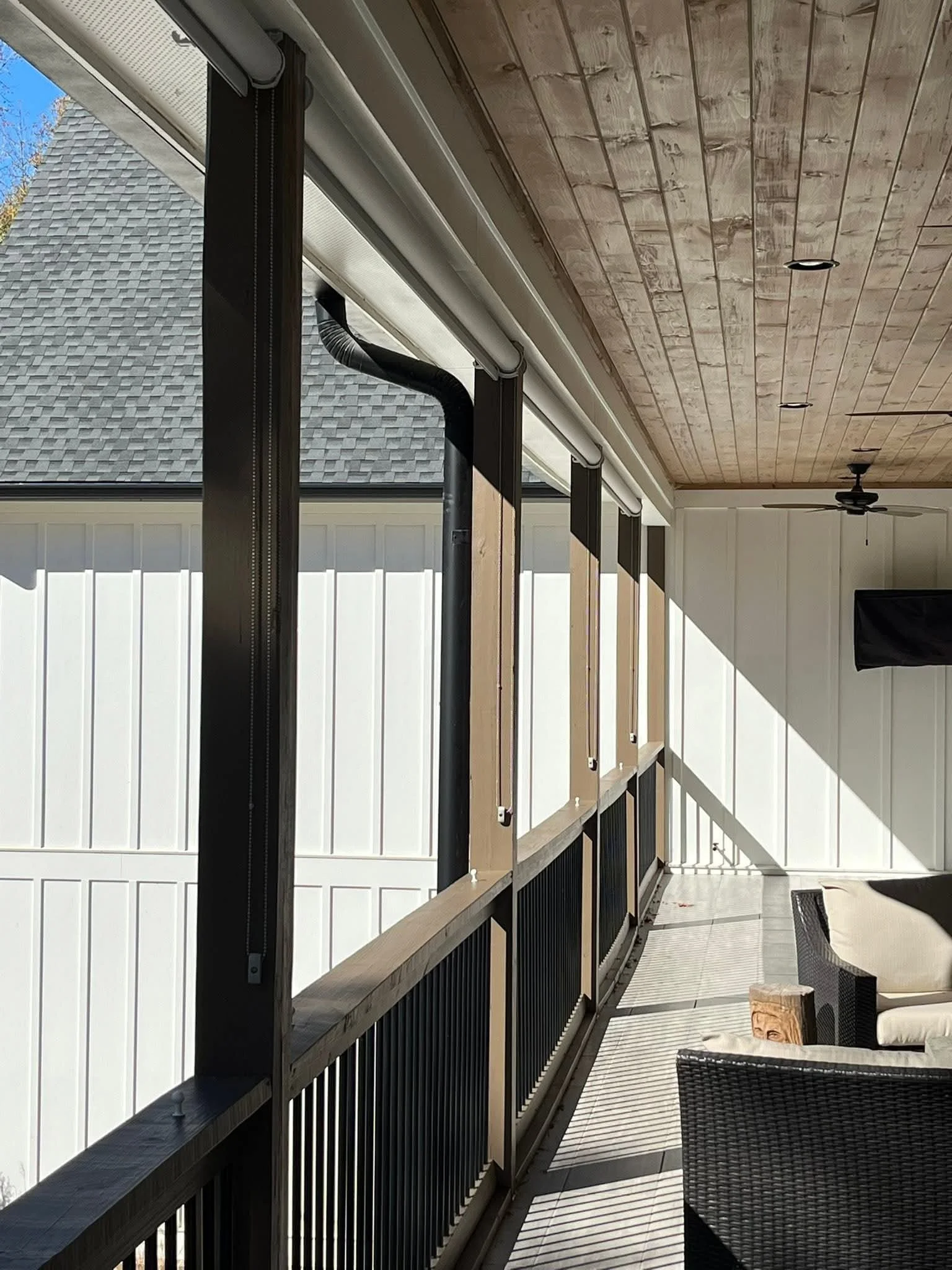 A porch or balcony area with a black railing, outdoor cushions, a wooden ceiling, and a ceiling fan. The space is partially enclosed with a white wall and has a view of a neighboring house's shingled roof and trees outside.