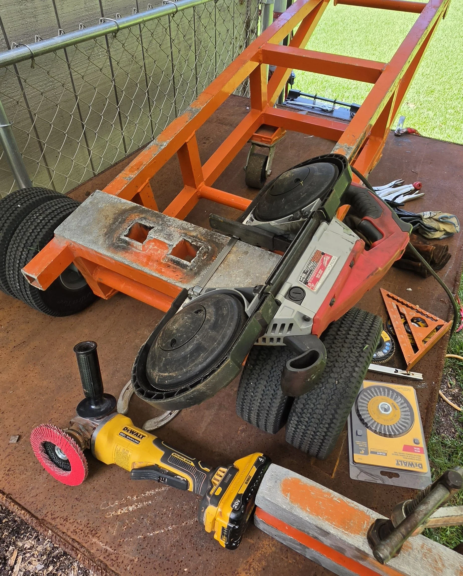 Workshop table with a partially assembled orange metal cart frame, power tools, sandpaper, and cleaning brushes, with a chain-link fence and grass in the background.