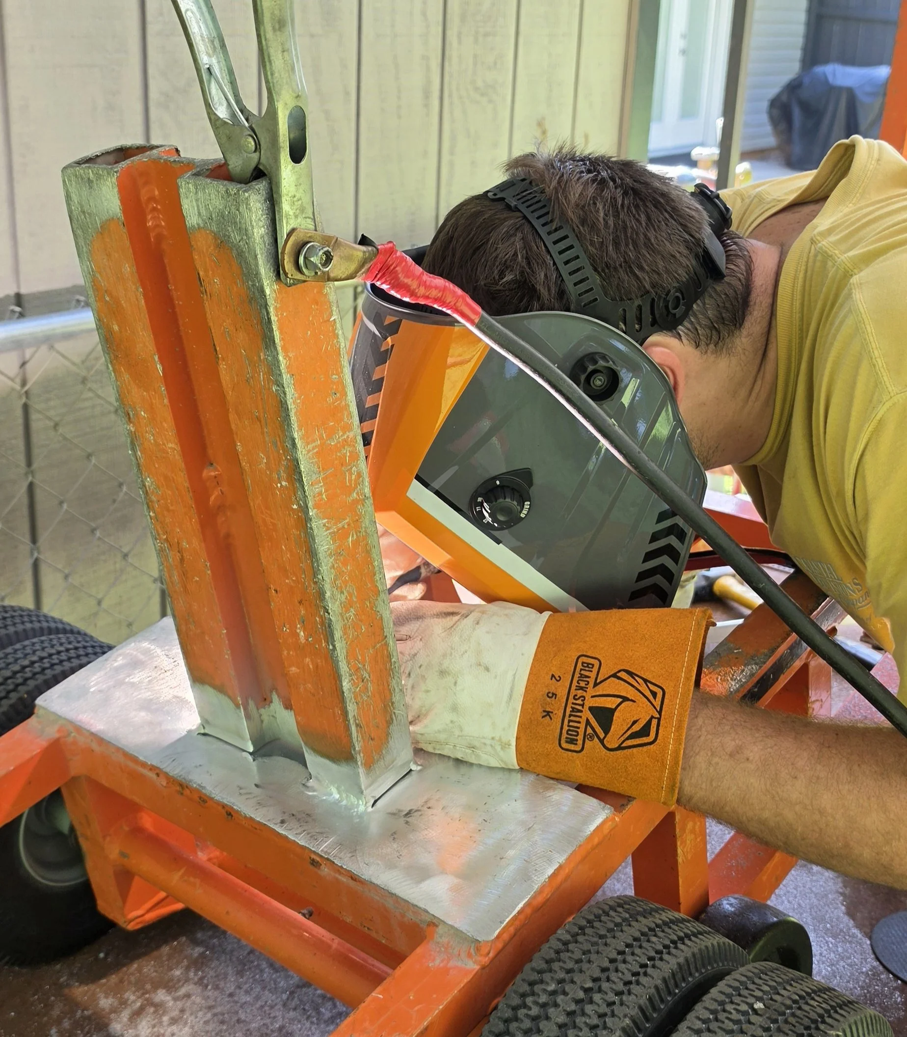 A person wearing a yellow shirt and safety goggles leaning over a workbench with an orange frame, working on a large piece of equipment connected with red electrical tape. The background shows a porch or outdoor space with siding and a door.