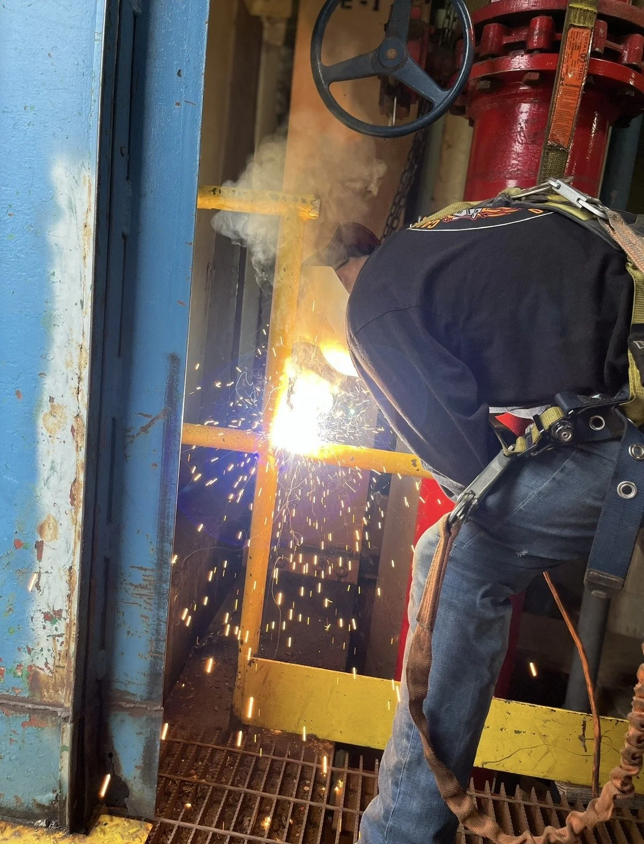 A worker is welding metal inside a structure, sparks and bright light are visible from the welding process, and the worker is wearing protective gear including a harness and helmet.