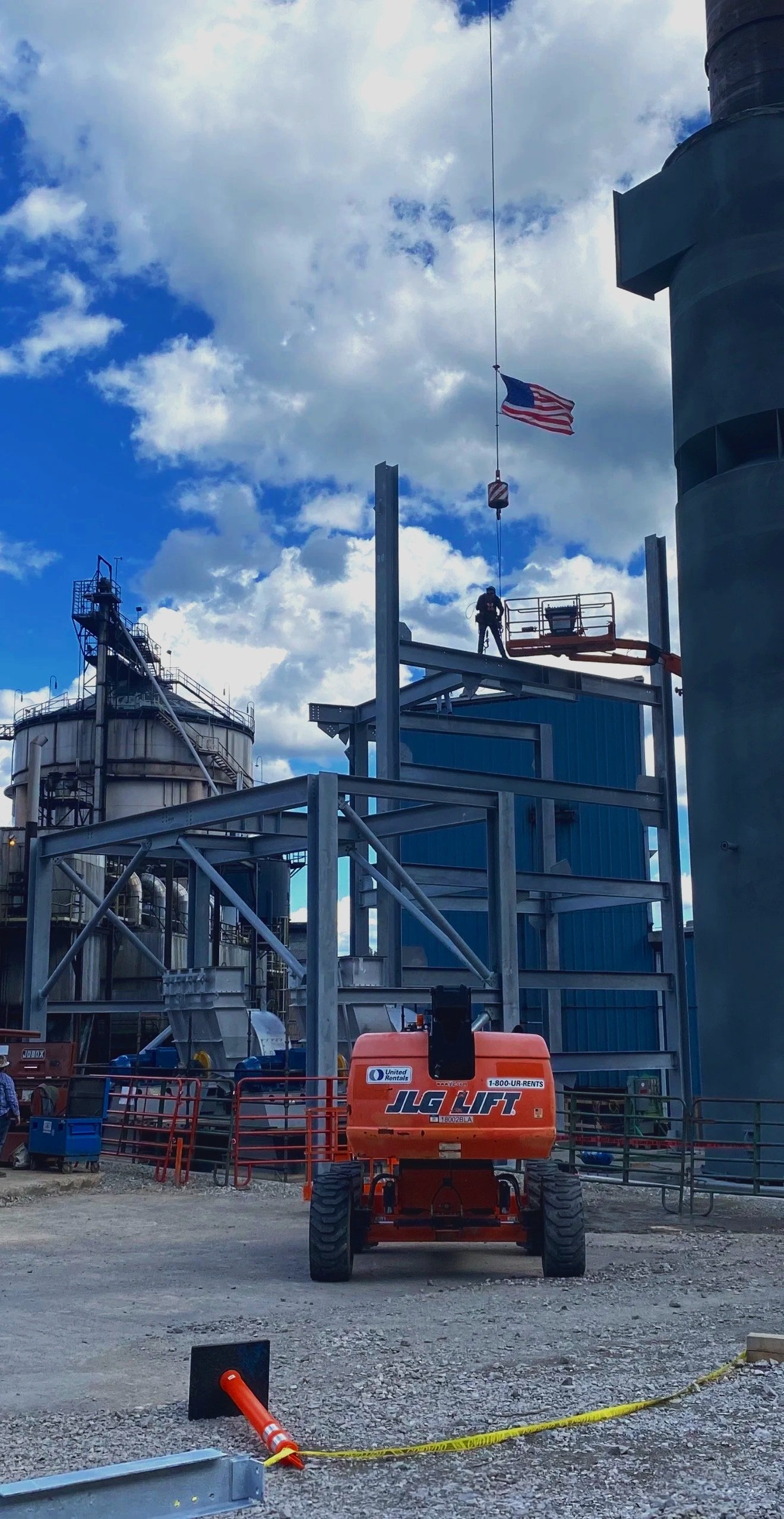 Construction site with a worker on elevated platform, American flag flying, and industrial structures in the background.