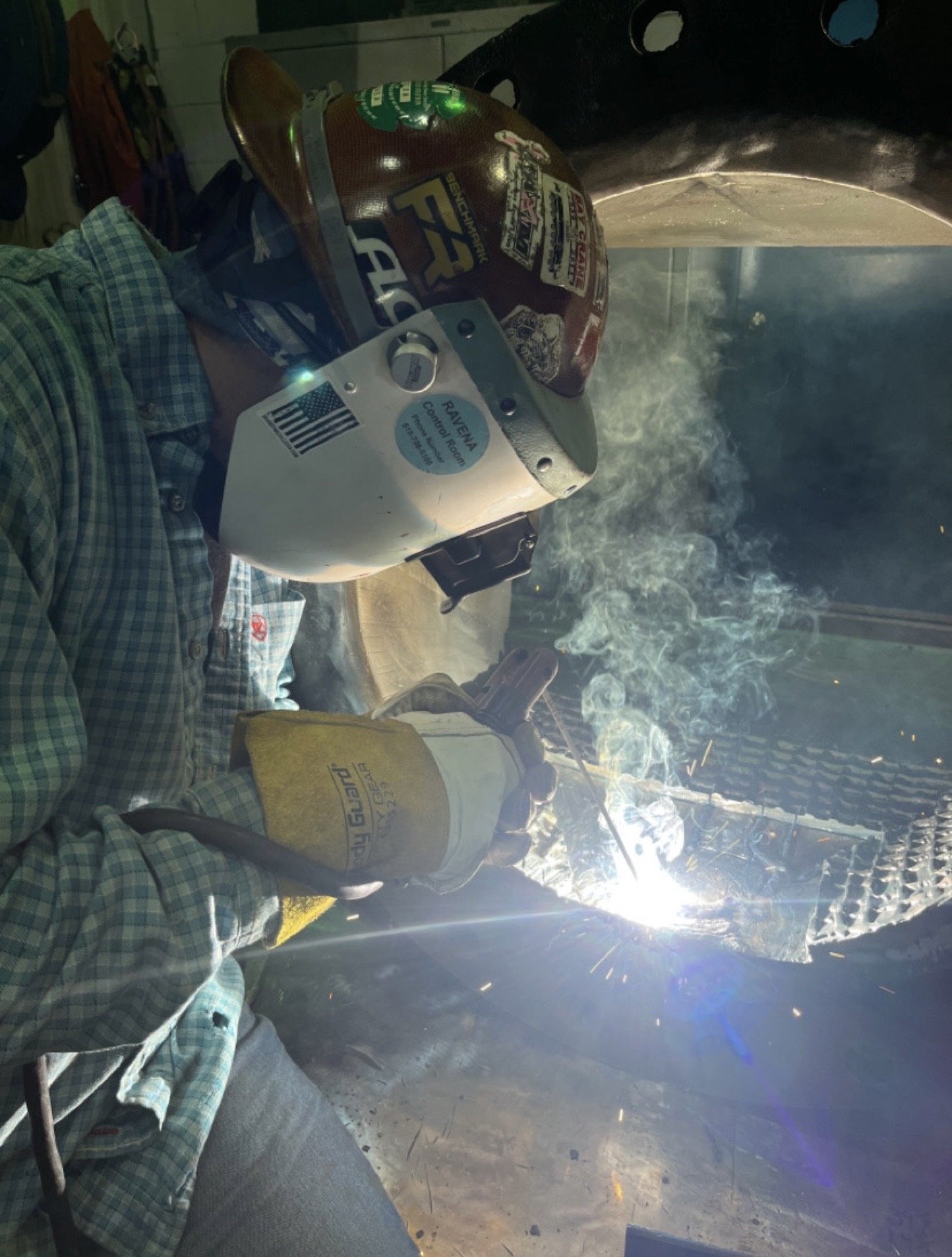 A welder wearing a plaid shirt, protective gloves, and a helmet with stickers, welding metal while sparks and smoke are visible, inside a workshop or garage.