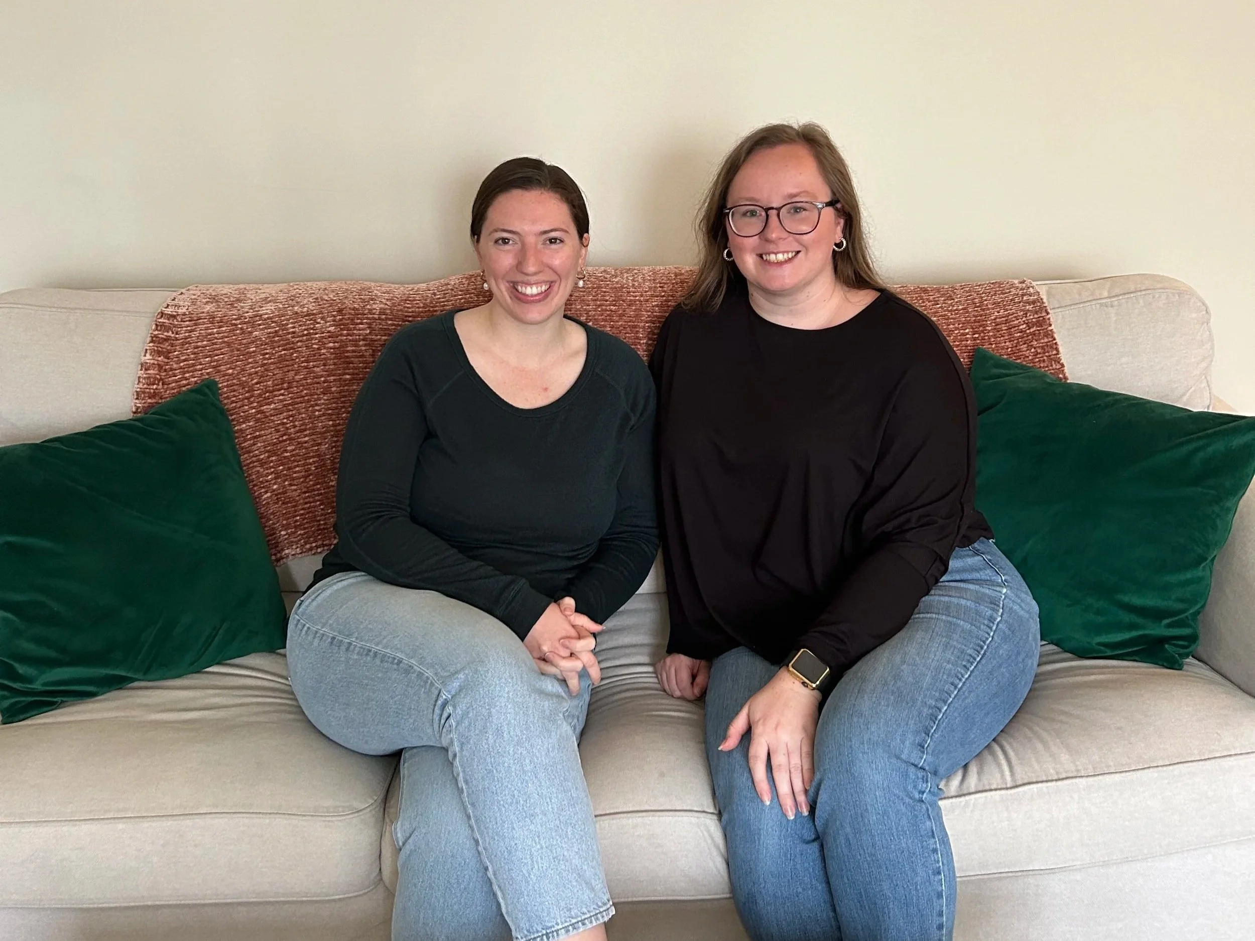 Two women sitting close together on a beige couch, smiling at the camera. They are surrounded by green pillows and a rust-colored throw blanket.