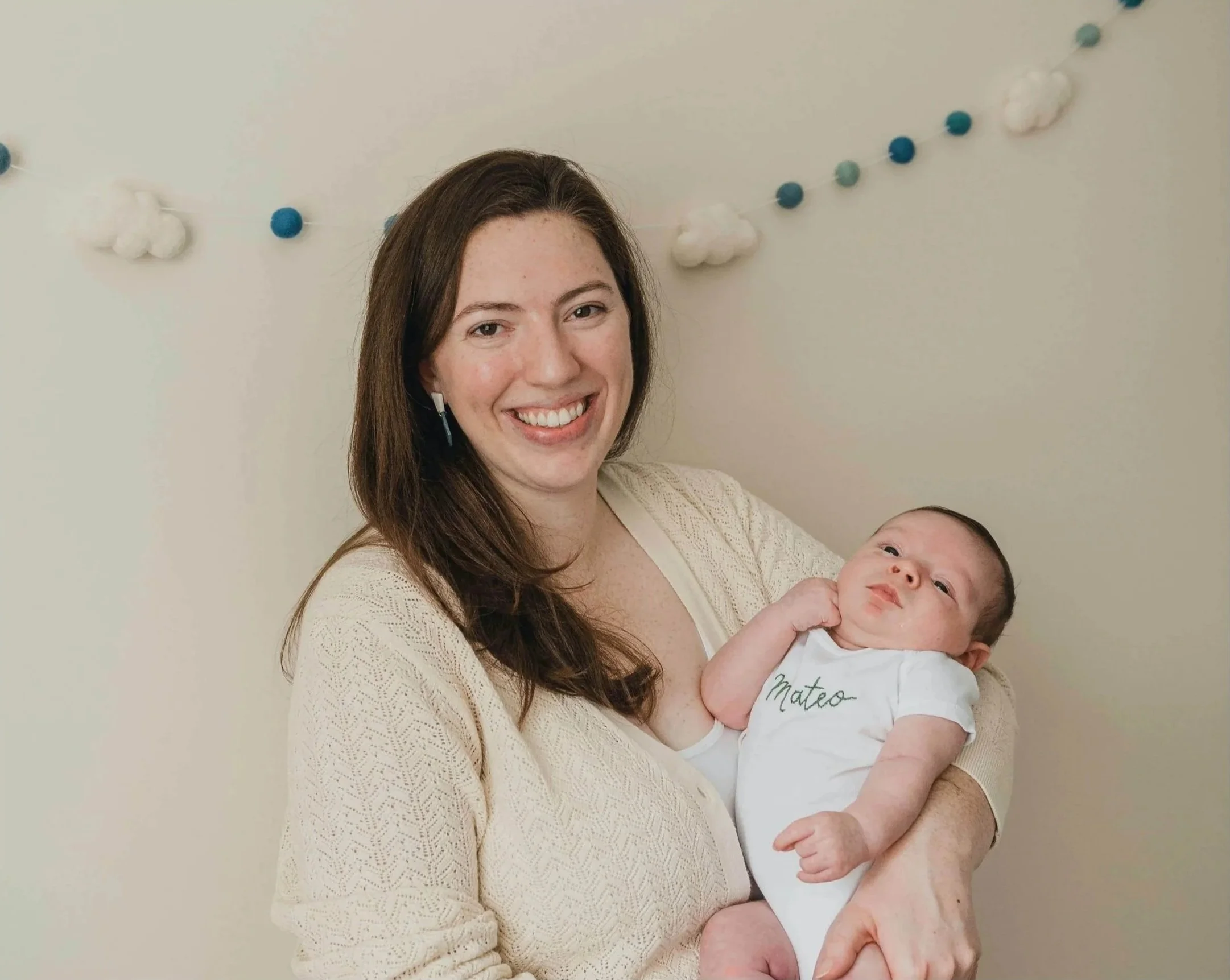 A smiling woman with long brown hair holds a newborn baby, who is wearing a white onesie with the name Mateo written on it, against a plain background with a decorative garland of cotton and felt balls hanging behind them.