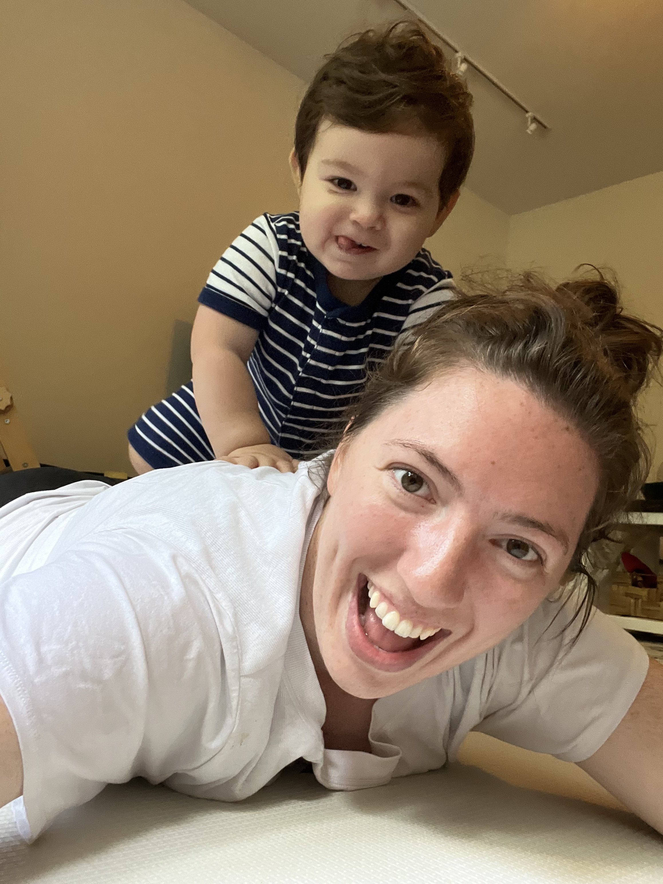 A woman with curly hair smiling and lying on the floor, with a young boy on her back, making a playful face. The boy has dark brown hair and is wearing a navy and white striped shirt.