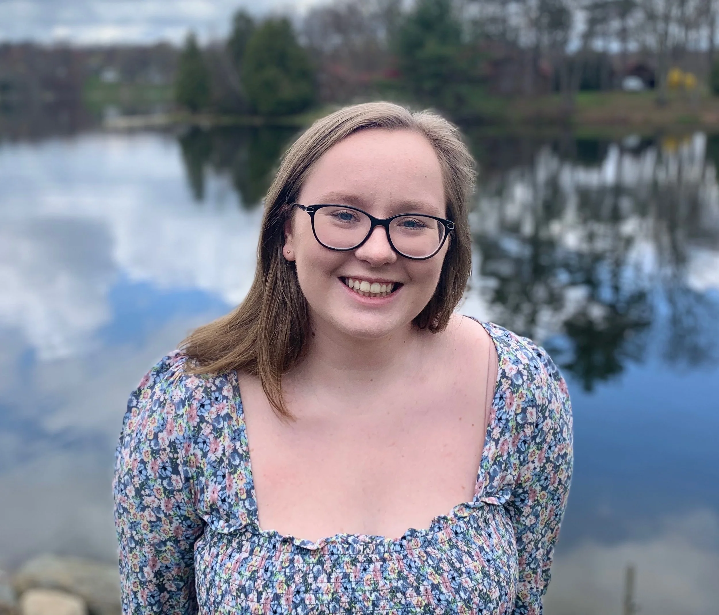 Close-up of a smiling young woman with light brown hair, wearing glasses and a floral dress, standing outdoors by a lake with trees and houses in the background.