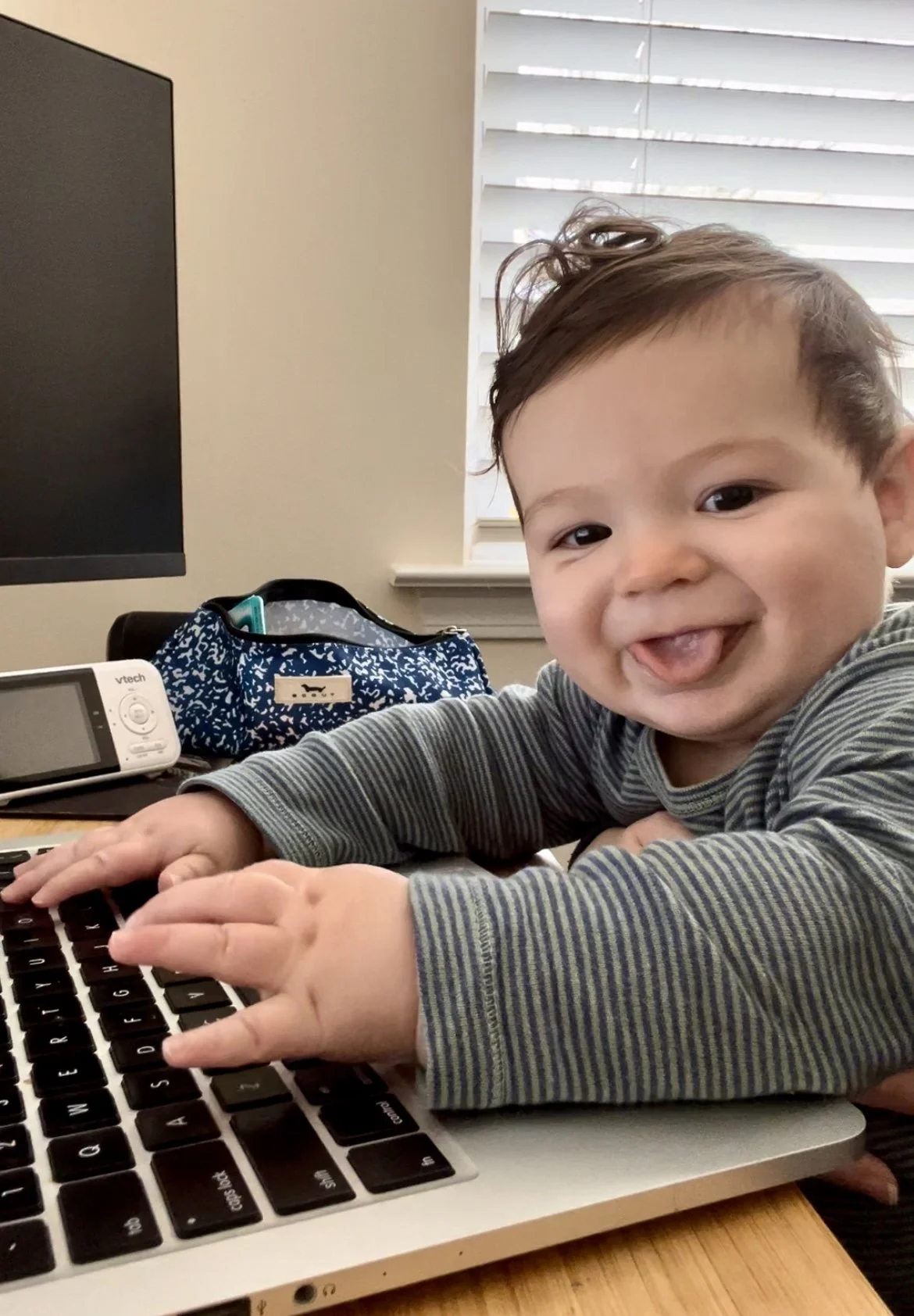 A smiling toddler with brown hair playing on a laptop keyboard in a room with a window and a blue patterned backpack on the desk.