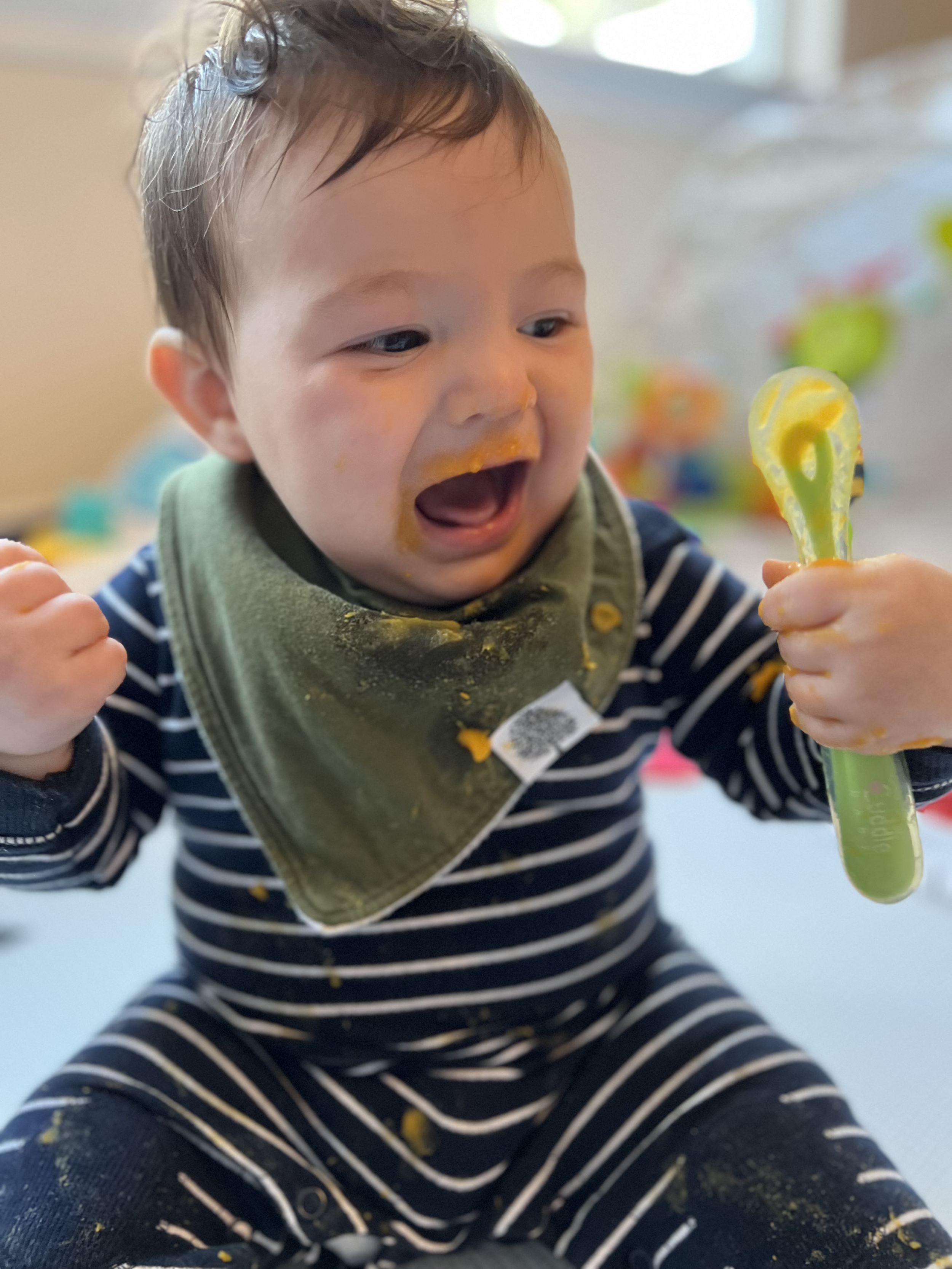 A young child with messy hair, wearing a striped shirt and a green bib, is happily playing with a yellow plastic spatula covered in orange baby food, with some food on the child's face and hands.