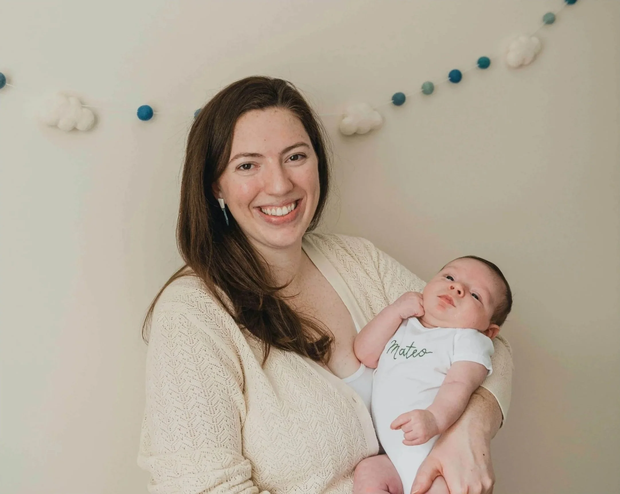 A smiling woman with long brown hair holding a baby with a light complexion and short dark hair. The woman is wearing a cream-colored sweater, and the baby is wearing a white onesie with the name 'Mateo' embroidered on it. There are decorative clouds and blue and white balls hanging on the wall behind them.