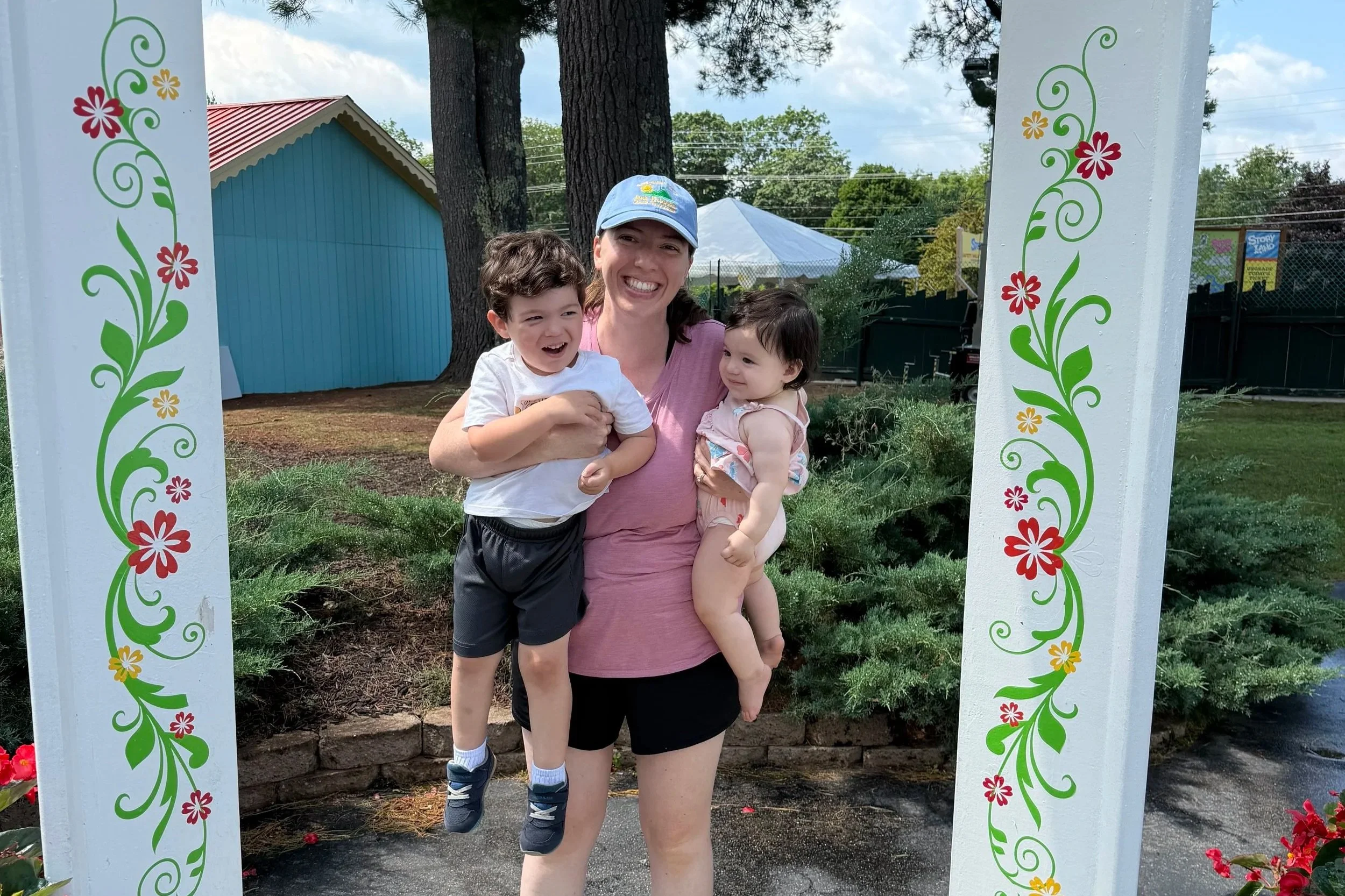 A woman holding a young boy and girl standing under a decorative white arch with red, yellow, and green floral designs in an outdoor park with trees and a building in the background.