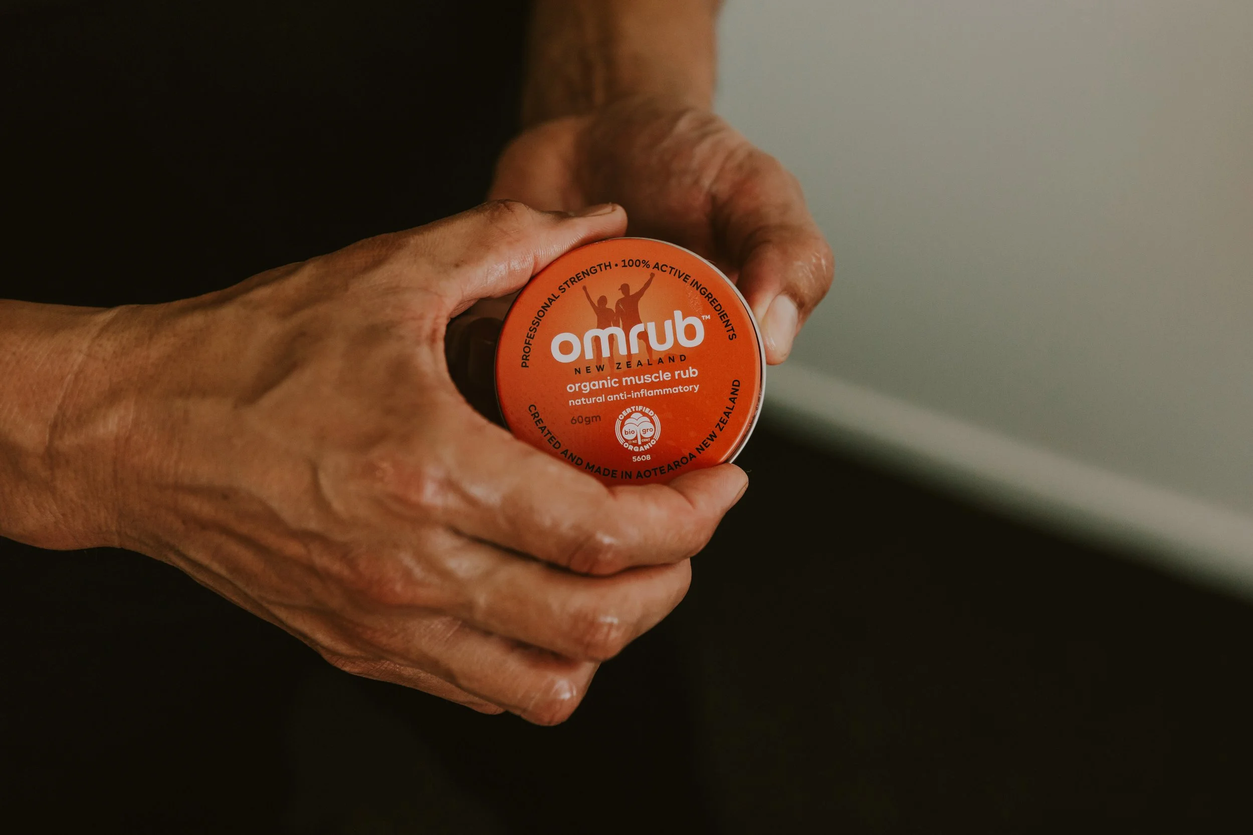 A person's hands holding a container of Omrub organic muscle rub with an orange label, against a neutral background.