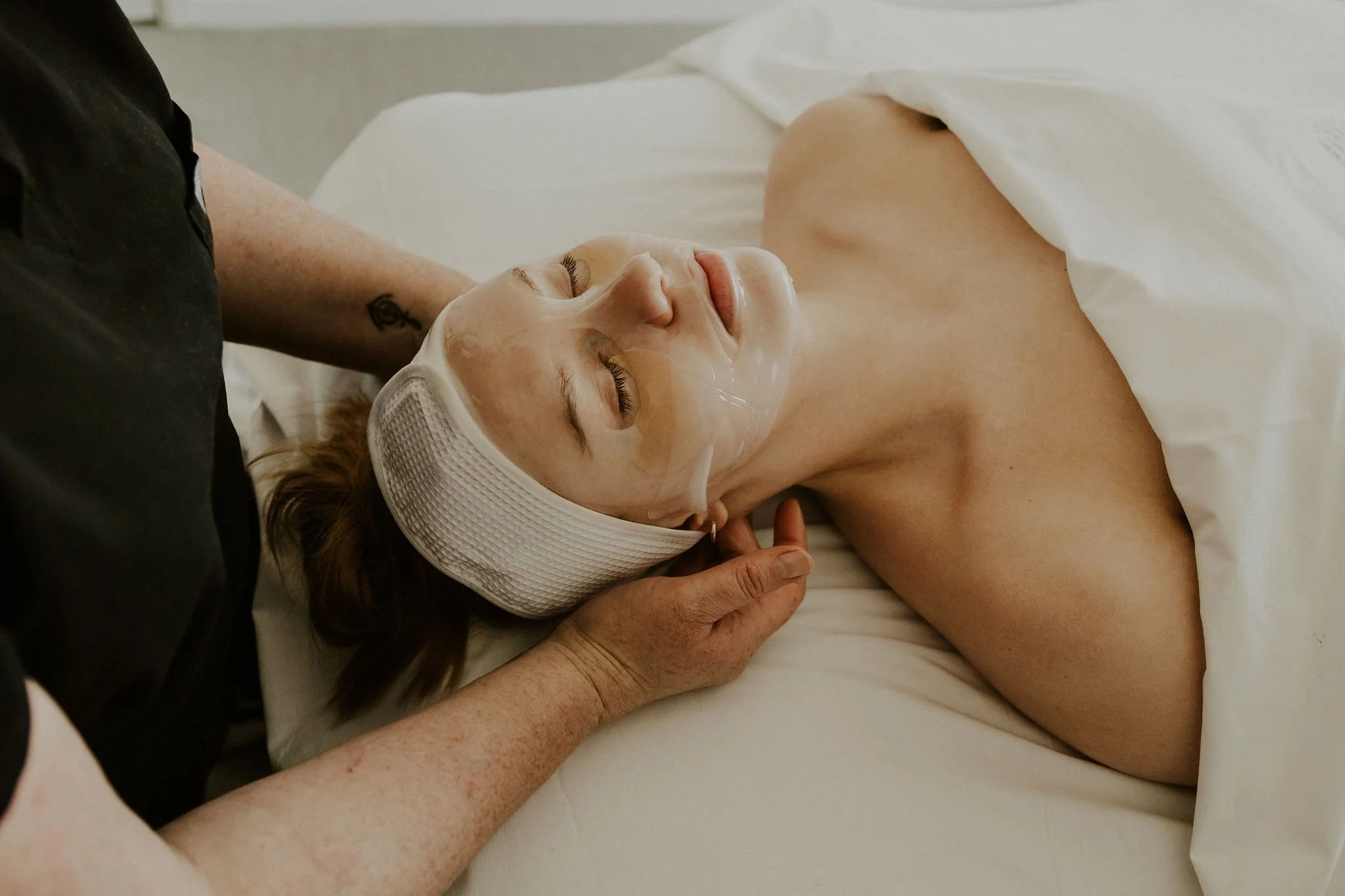A woman lying on a bed with her eyes closed, receiving a facial treatment, wearing a headband, with a white sheet covering her body, and a person standing near her with a tattoo visible on their arm.