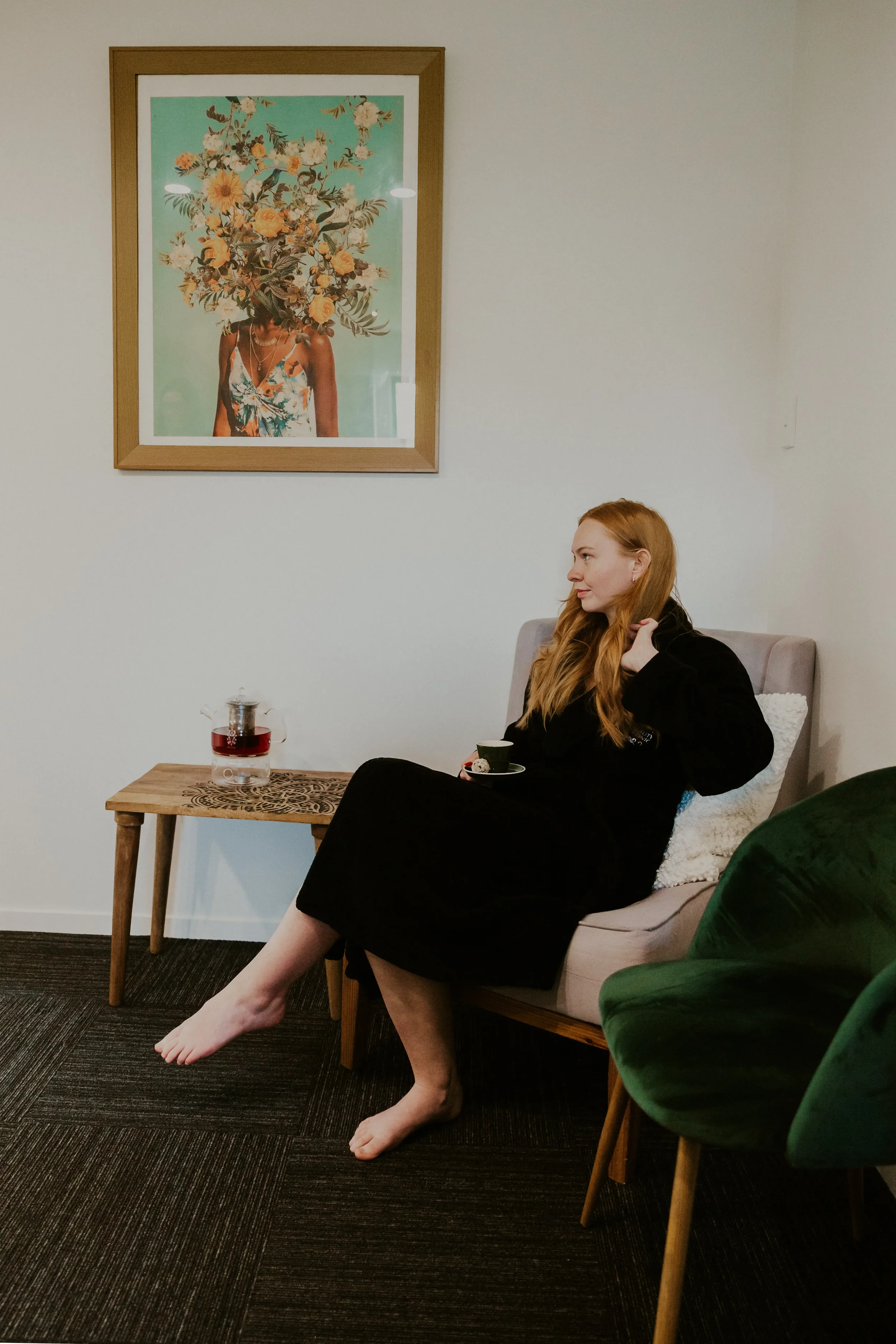A woman with long red hair sitting on a beige couch, barefoot, holding a black mug, wearing a black dress, in a room with a framed artwork of a woman with a bouquet as her head on the wall. There is a small wooden table with a glass container and a red drink in front of her, and a large green plant in the foreground.
