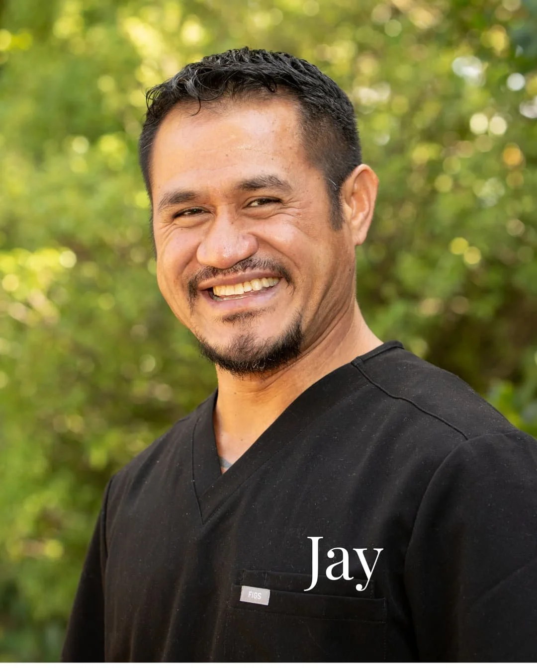 A smiling man with short dark hair and a goatee, wearing a black medical uniform with the name tag 'FIGS', standing outdoors with greenery in the background.