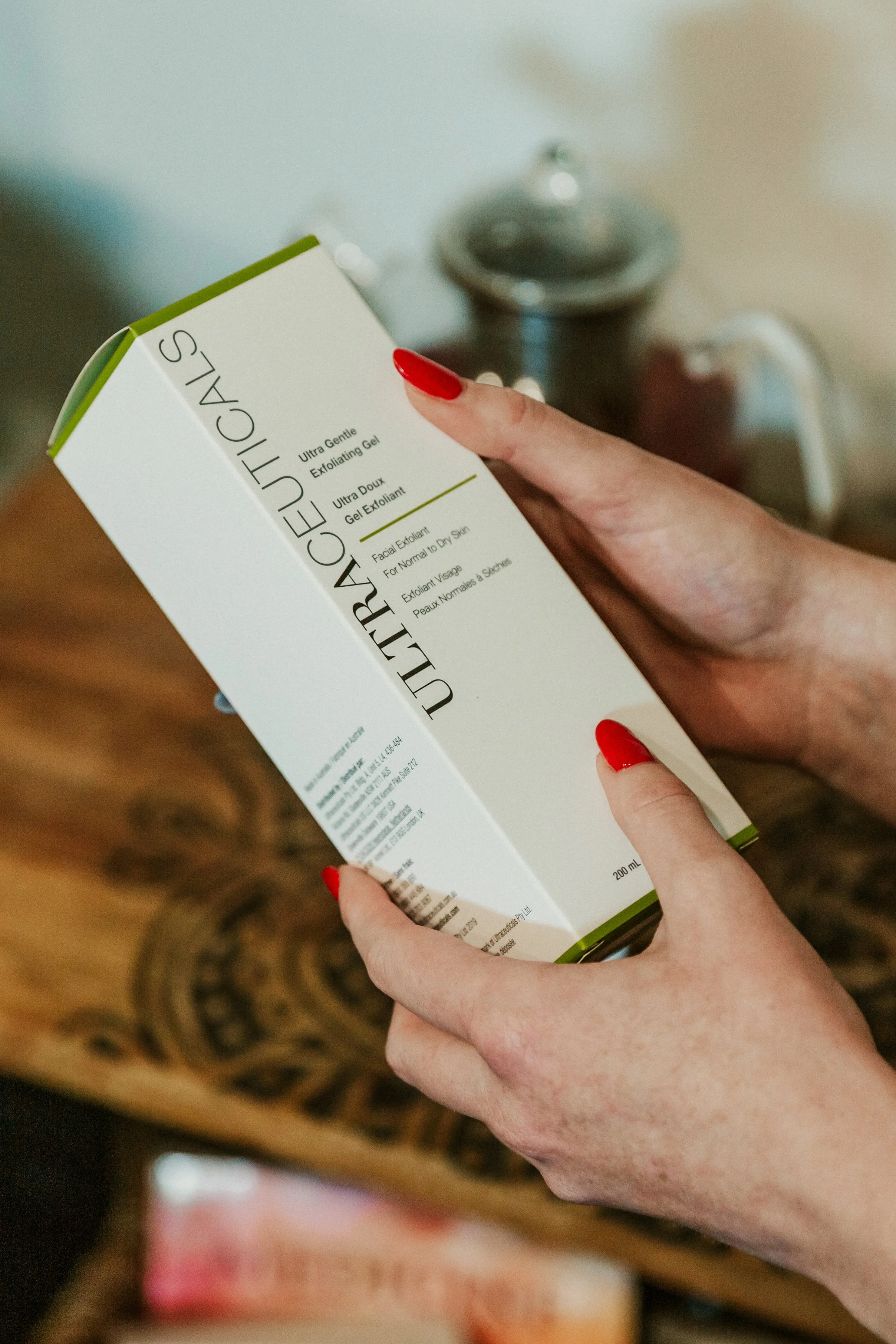 A person's hands holding a box of luxurious exfoliating gel with red nail polish. In the background, a water kettle and a mug are visible on a wooden surface.