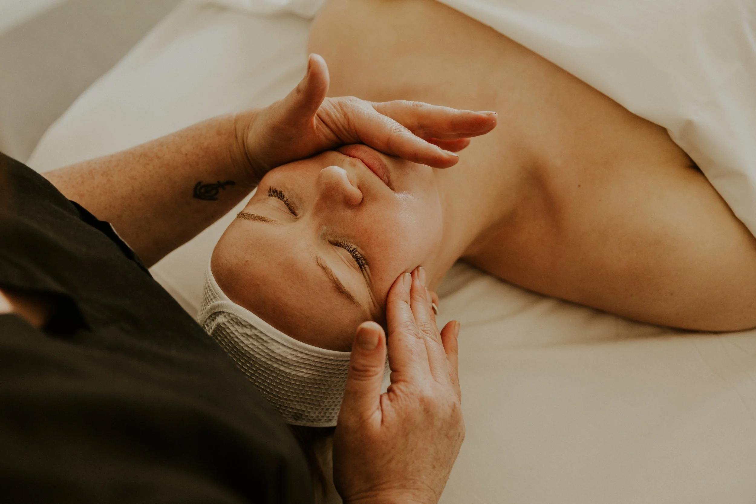 Person receiving a facial massage in a spa setting, lying on a bed with a white sheet, with a hand gently touching their forehead.