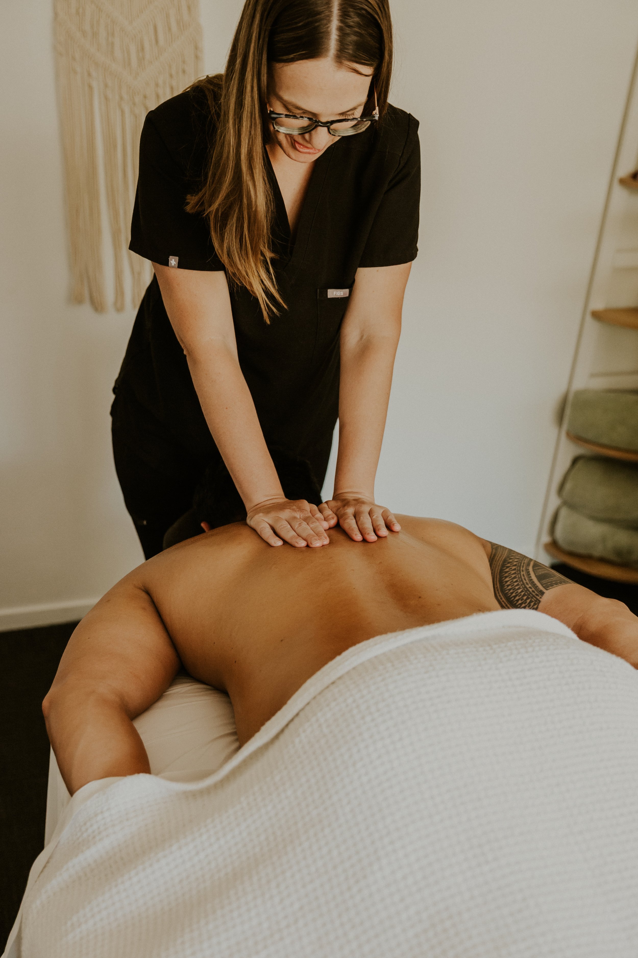 A chiropractor giving a massage to a man lying face down on a massage table, with a towel covering his lower body and a tattoo on his left arm.