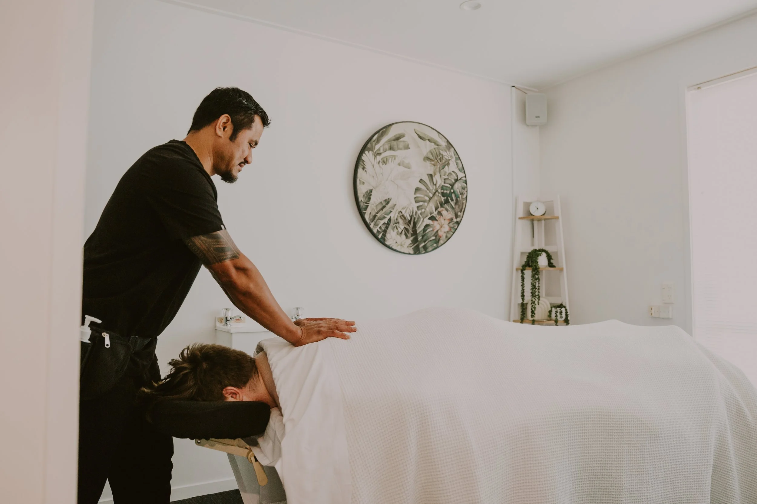 A person receiving a massage in a bright, minimalist room with tropical decor art on the wall and a small shelf with a clock and plants.