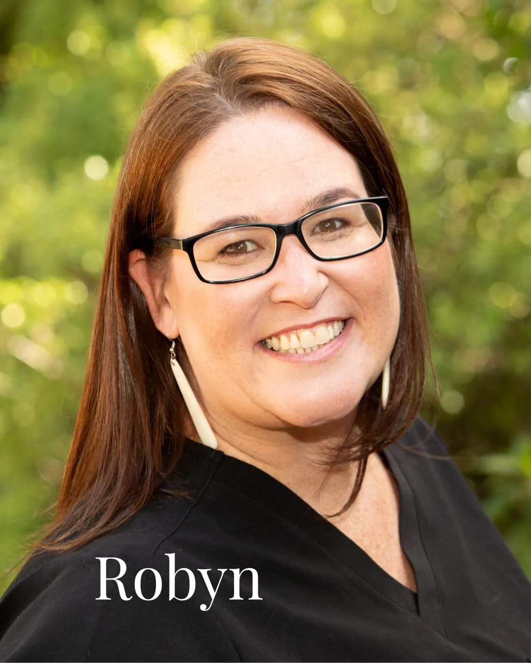 Close-up of a woman with brown hair, glasses, and earrings, smiling outdoors with a blurred green background. The name 'Robyn' is written at the bottom.