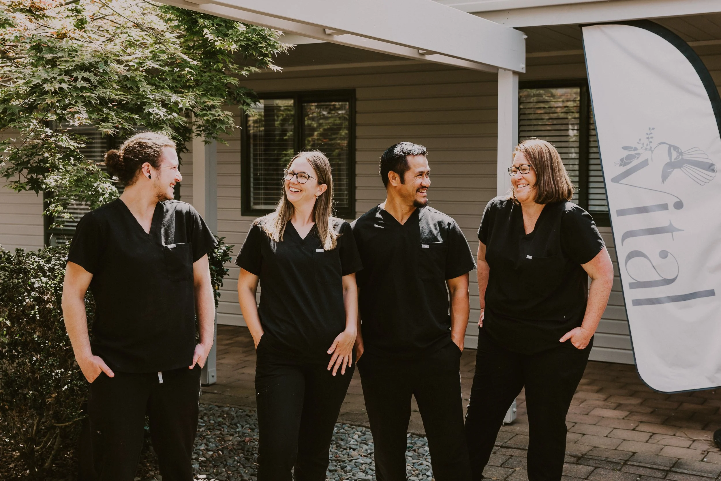 Four people wearing black scrubs standing outdoors, smiling and talking to each other, with a house and a banner in the background.